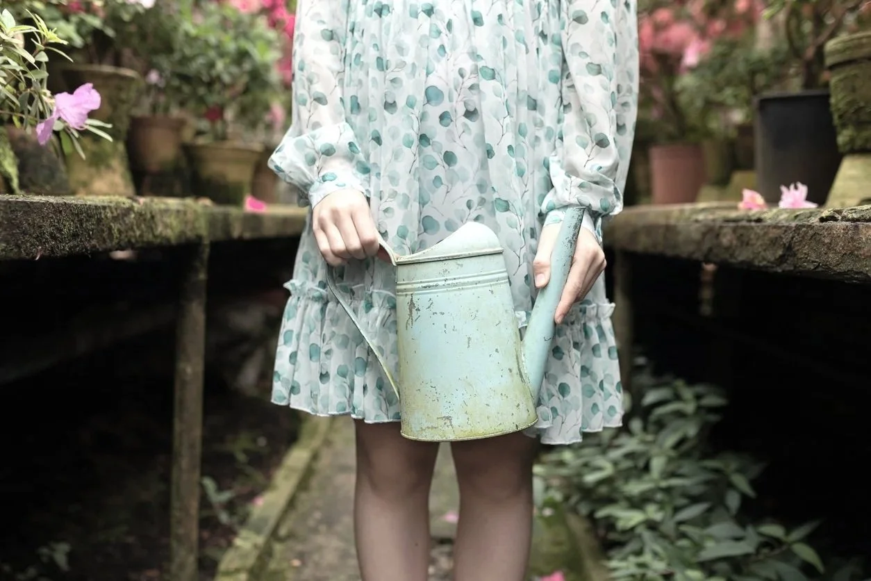 A person in a floral dress watering plants in a garden or greenhouse.