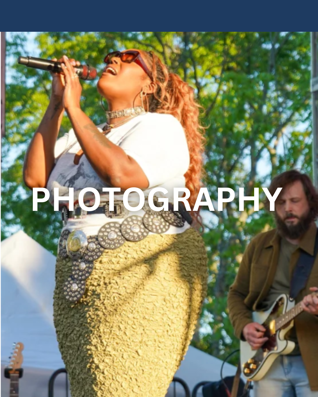 A woman singing into a microphone during an outdoor performance with a guitarist playing beside her, trees and a tent in the background.