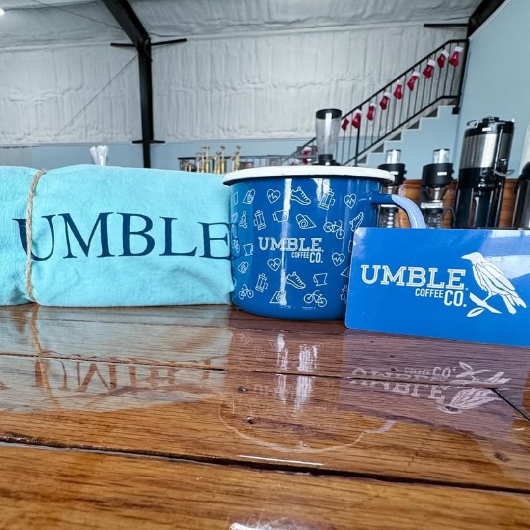 Blue coffee mug with the "UMBLE COFFEE CO." logo, a blue card with the same logo and a bird, and a light blue cloth with the word "UMBLE" on it, all placed on a wooden table inside a coffee shop.
