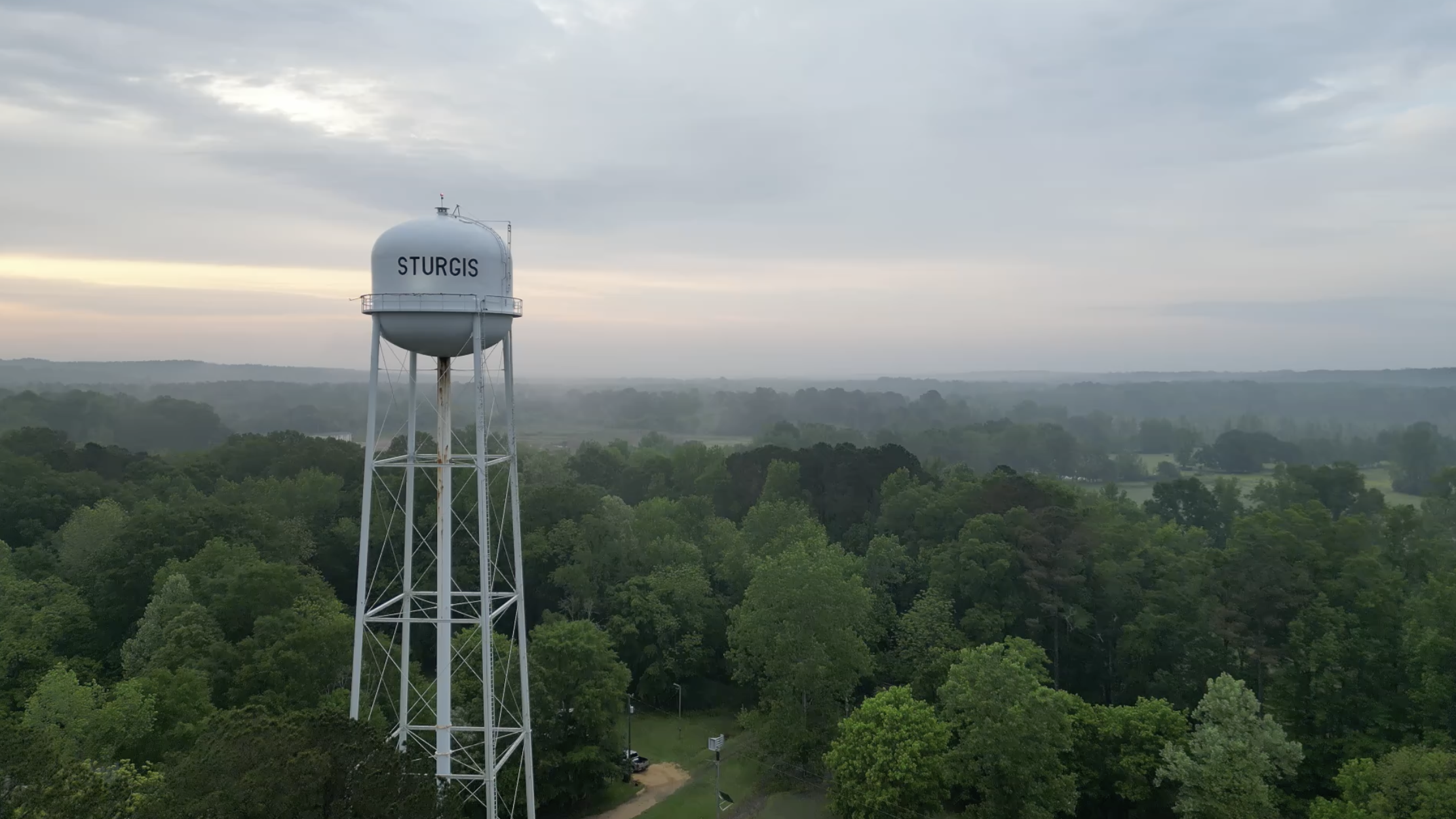 Water tower labeled 'STURGIS' standing above a dense green forest under a cloudy sky.