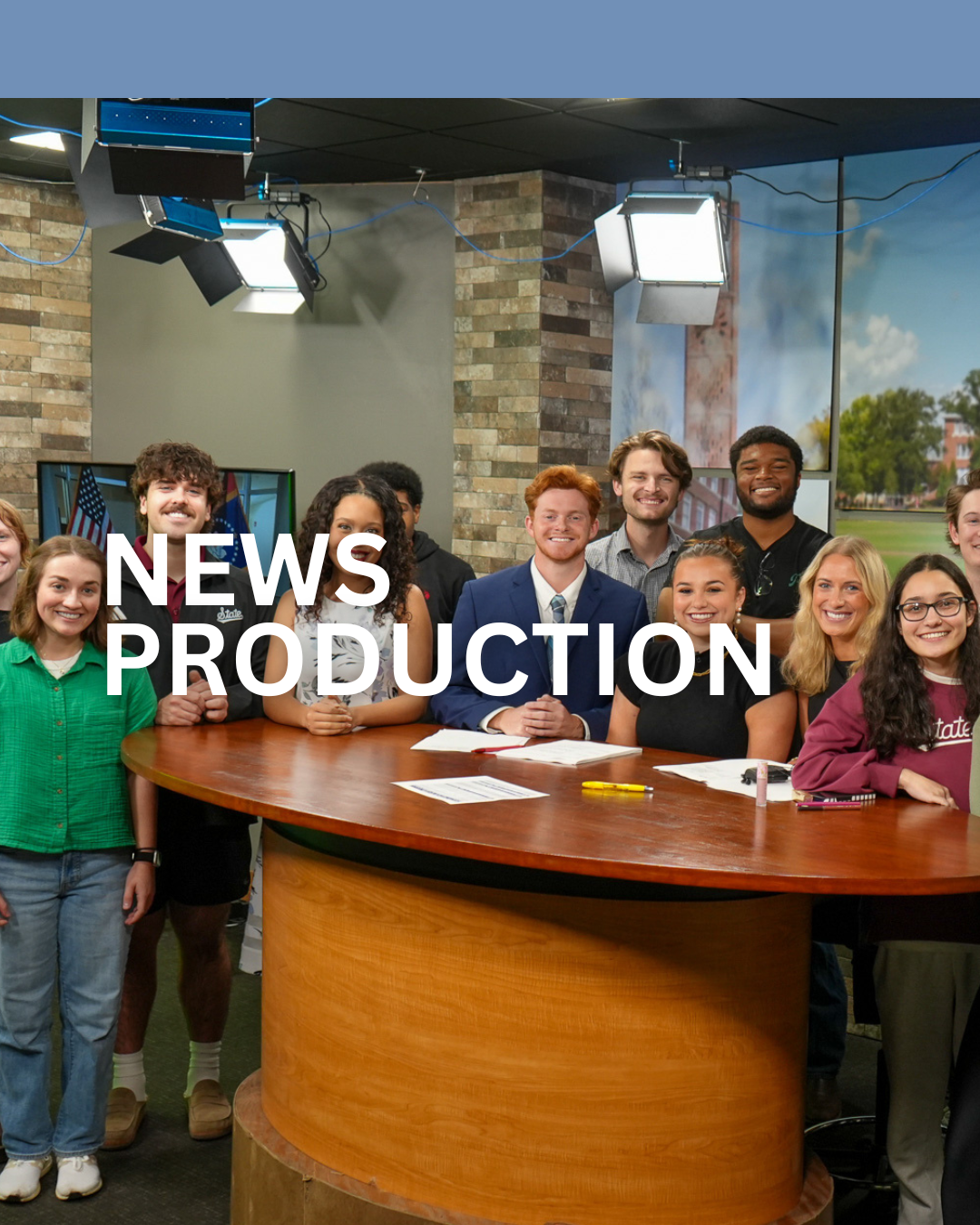 A group of diverse young people smiling behind a news desk in a television studio with equipment and large windows showing an outdoor field.