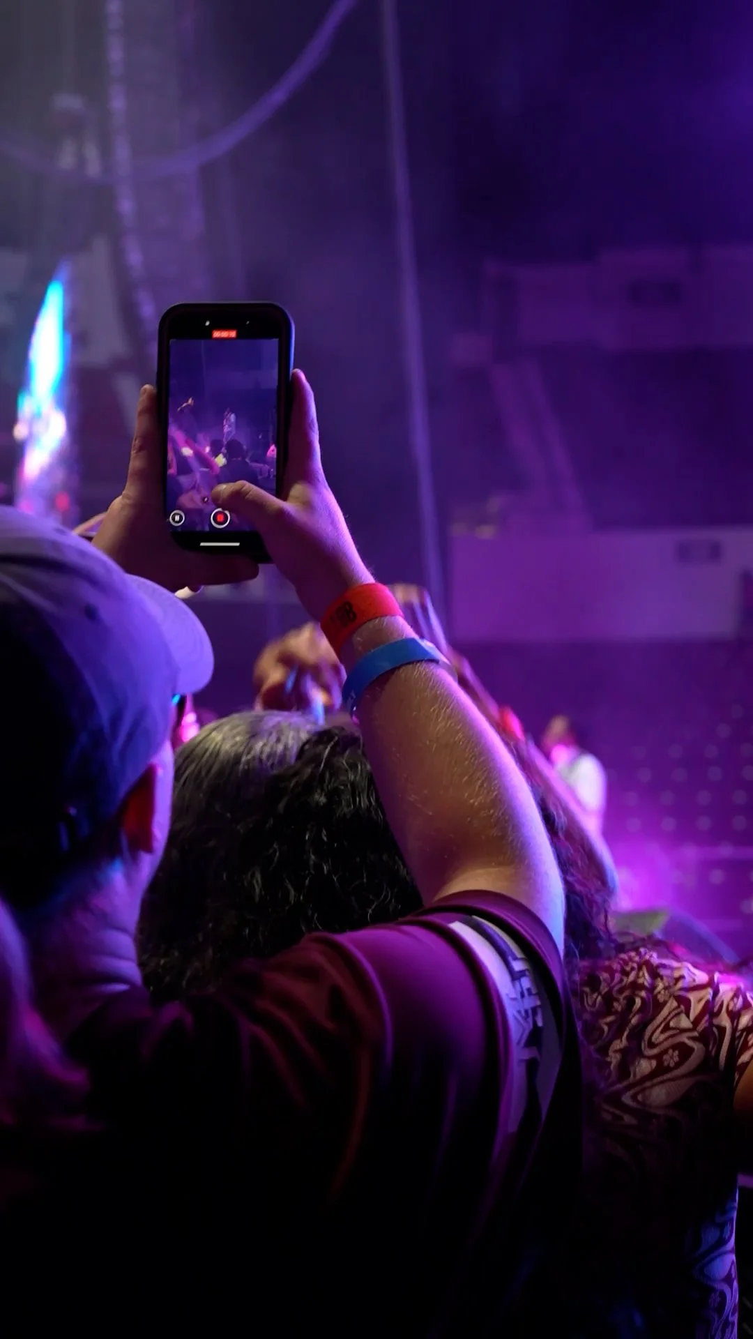 A person is taking a photo or recording a video with a smartphone at a concert or performance, with other people nearby. The scene is lit with purple and blue stage lights.