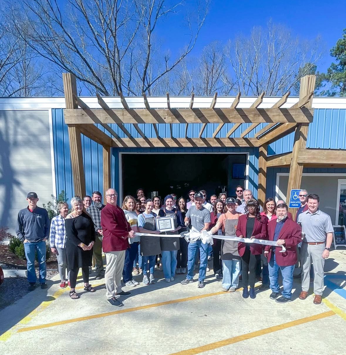 Group of people attending a ribbon-cutting ceremony for a new building with a wooden trellis and blue and white exterior walls.