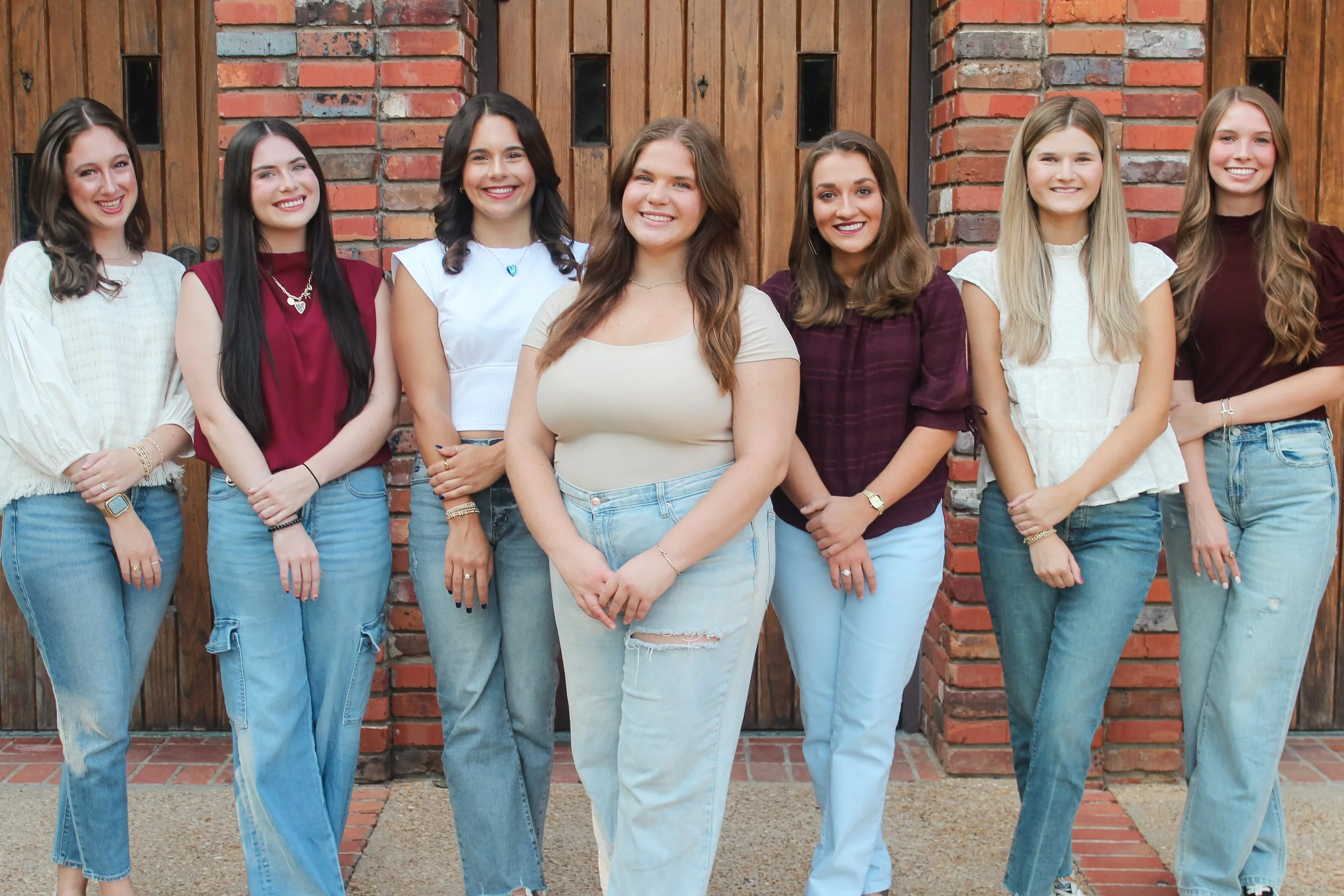 Group of eight young women standing together in front of a wooden and brick wall, smiling for the camera.