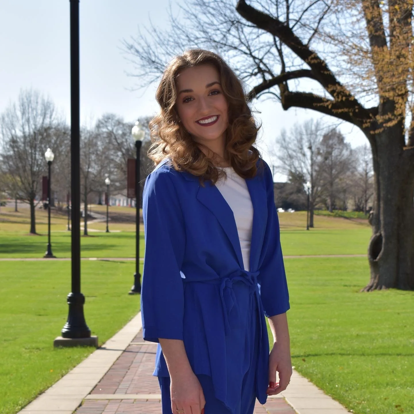 A young woman with curly brown hair, wearing a blue graduation gown and white shirt, standing outside on a campus-like area with green grass, trees, and lamp posts.