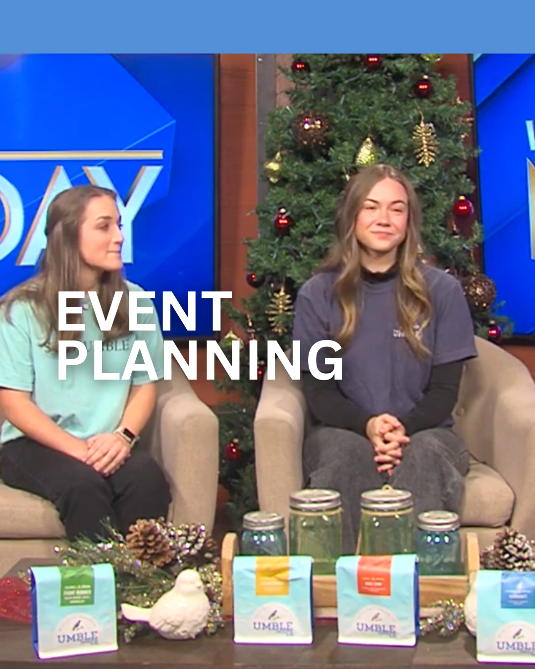 Two women sitting on sofas with a Christmas tree decorated with red and gold ornaments behind them, and a display of mason jars, pinecones, and tea packages on the table in front, during a segment about event planning.