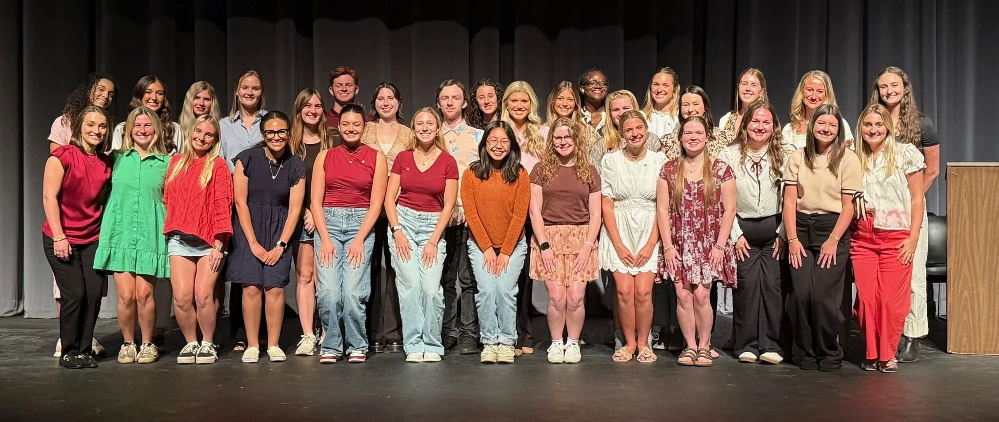 Group of young women and girls standing on stage for a photo, smiling, with a dark curtain backdrop and a wooden podium on the right.