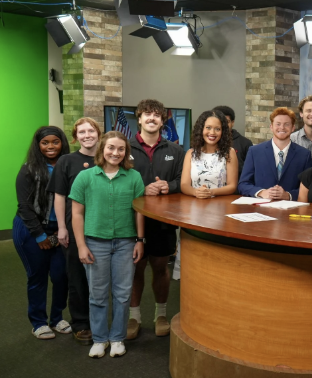 Group of seven young people, five women and two men, inside a television studio with a curved news desk, green screen, brick wall, and studio lighting.