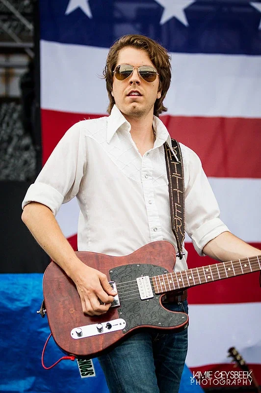 A man wearing sunglasses and a white shirt playing an electric guitar on stage with an American flag in the background.