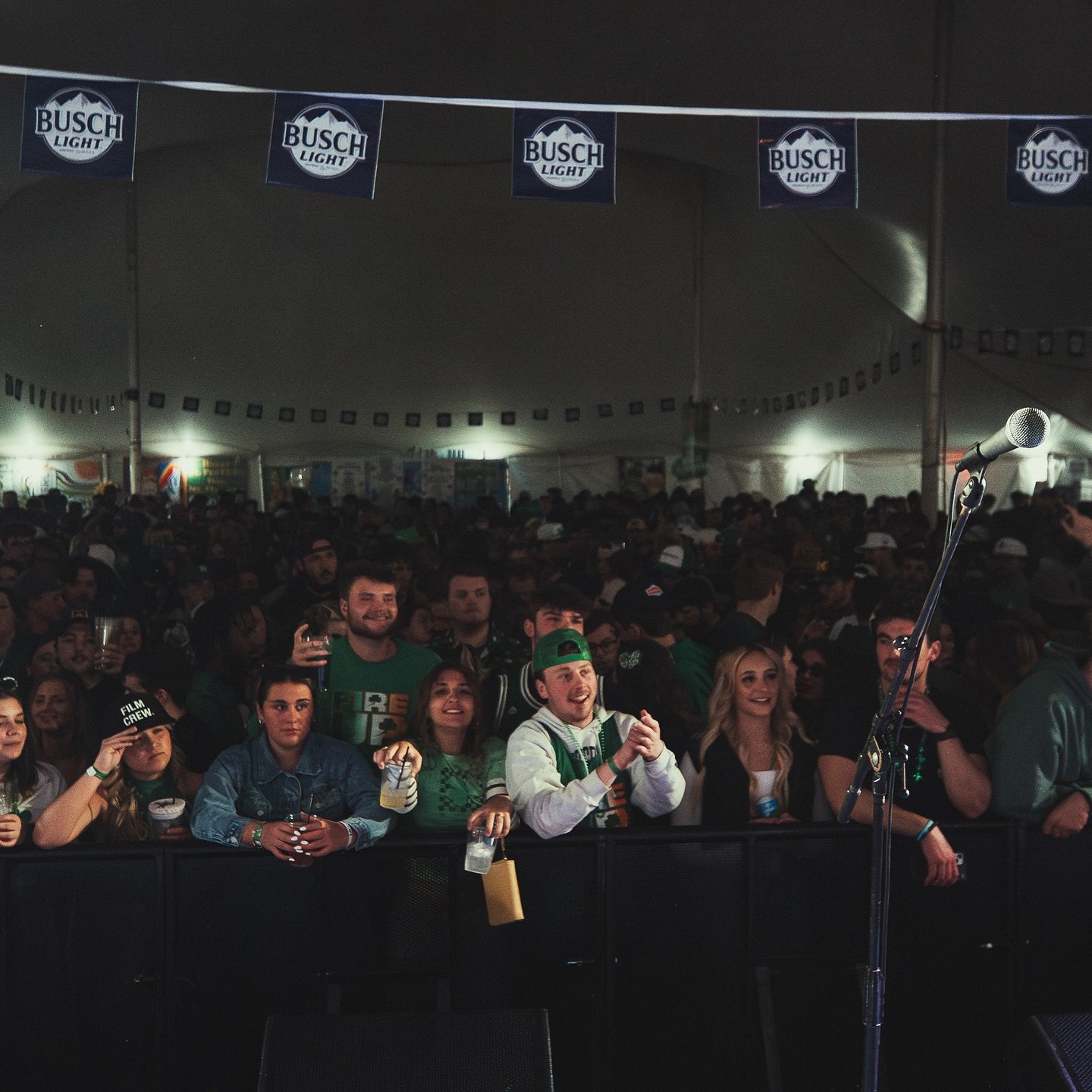 Crowd of people inside a large tent, some holding drinks, with a microphone stand in the foreground and banners for Busch Light hanging overhead.