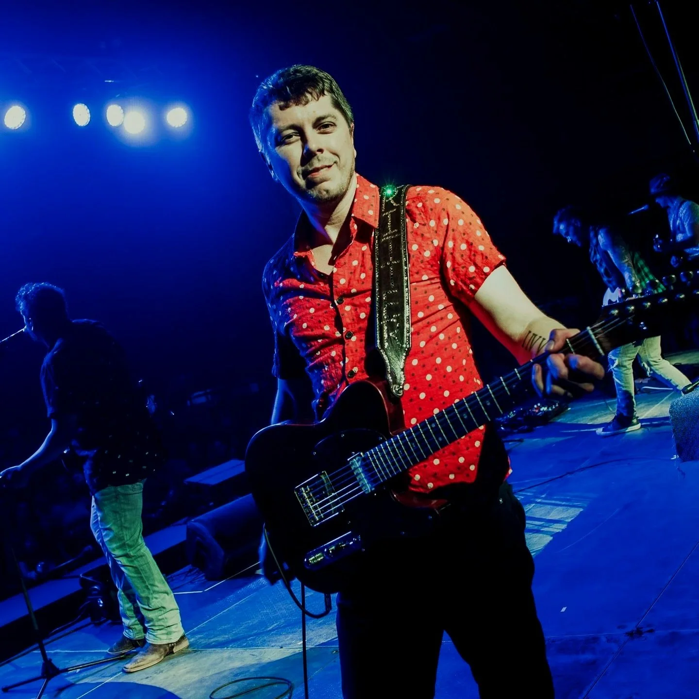 Musician in red polka dot shirt playing electric guitar on stage during a concert, with band members in the background and stage lighting.