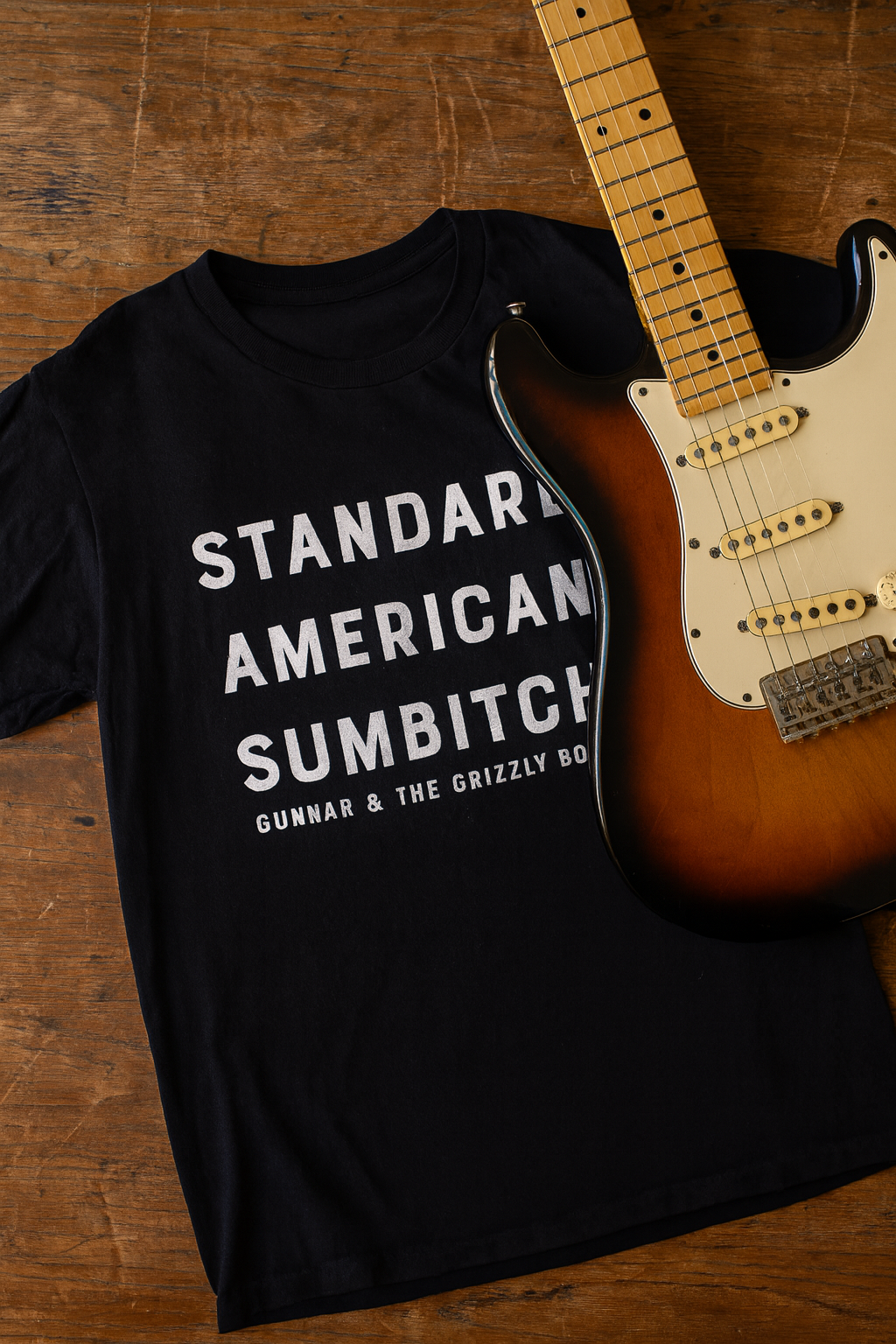 Black T-shirt with white text that reads 'Standard American Sambitch' and 'Gunnar & The Grizzly Boys,' laid on a wooden surface next to an electric guitar with a sunburst finish.