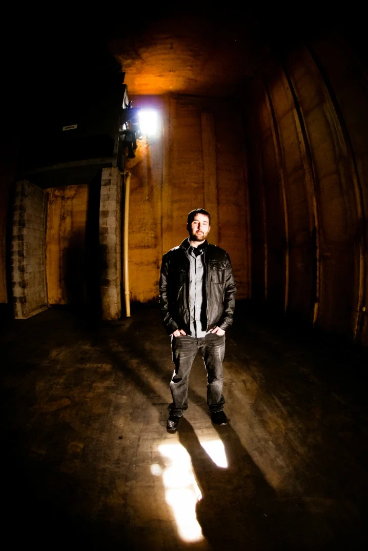 A man standing in a dimly lit wooden room with a high ceiling, illuminated by a bright light source behind him.