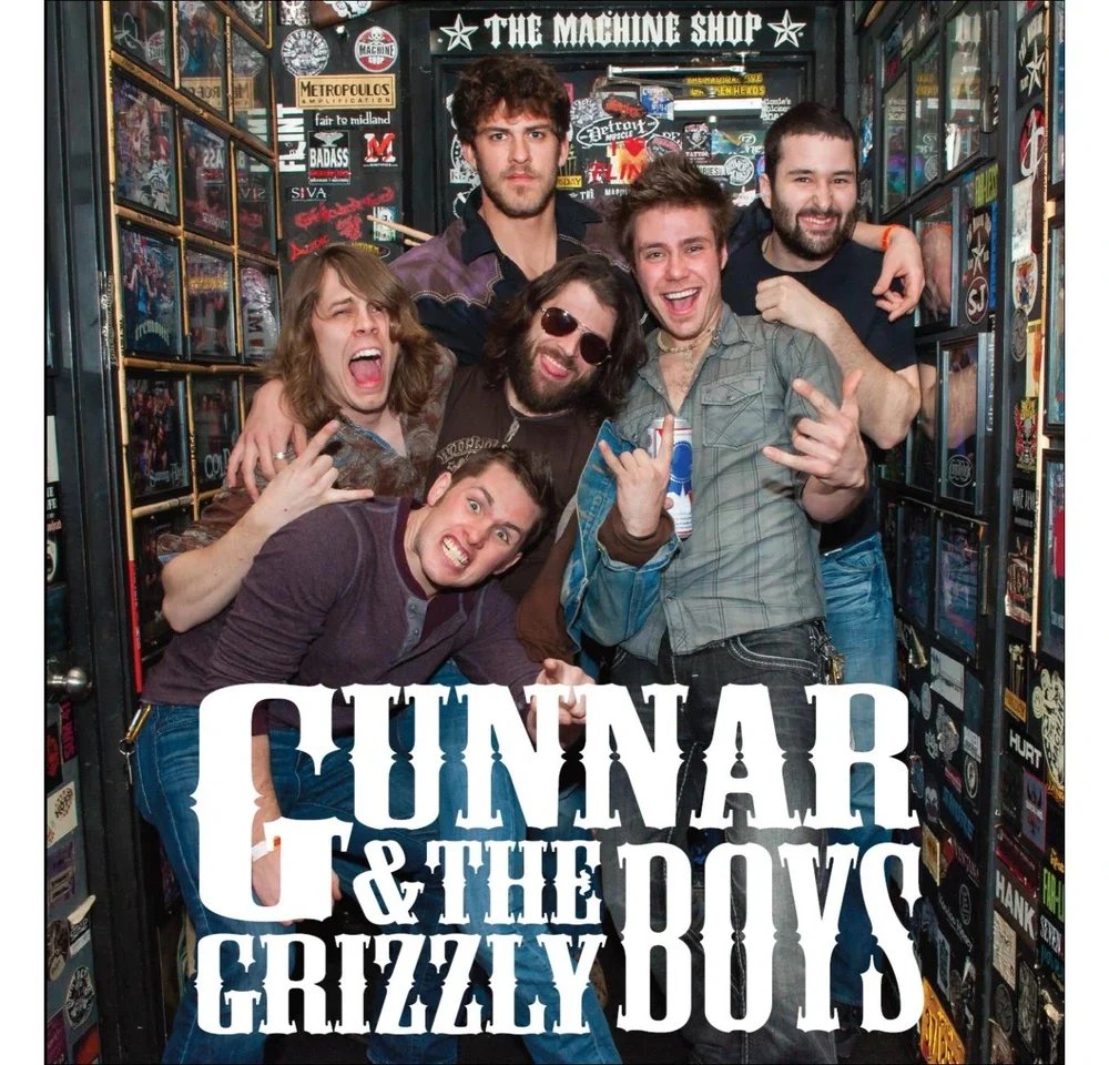 Six men posing in a small, sticker-covered room with a sign that reads 'The Machine Shop.' They are making rock and roll gestures and smiling at the camera, with one holding a beer.