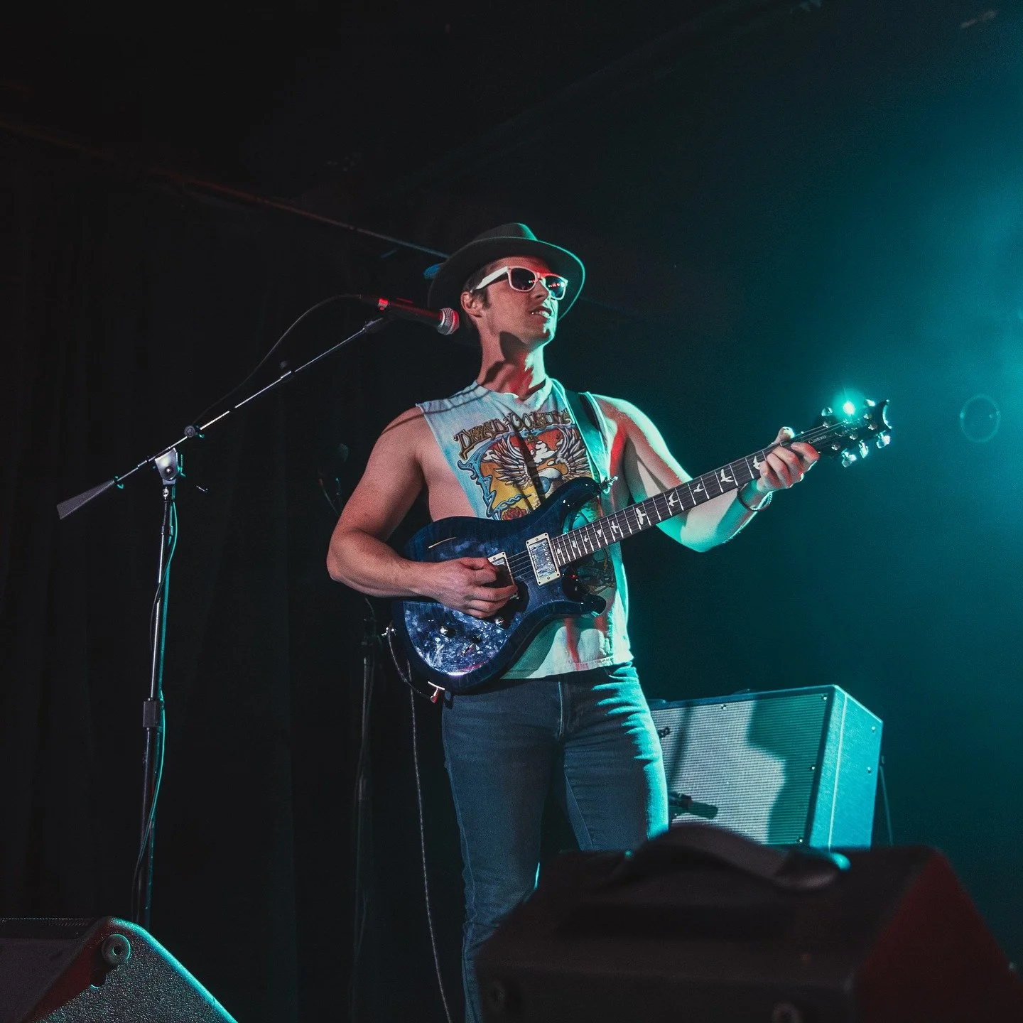 A musician on stage playing an electric guitar, wearing a sleeveless shirt, sunglasses, and a hat, with stage lighting illuminating the scene.