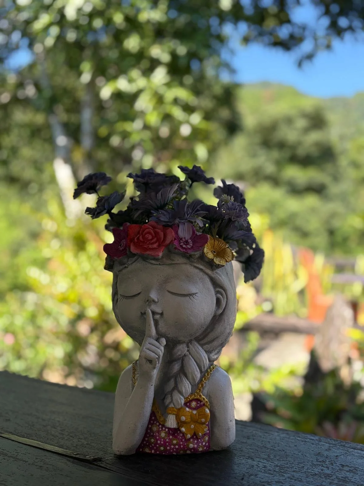 Sculture en poterie d'une jeune femme avec une tresse, portant un collier avec pendentif, qui fait un signe de silence en mettant un doigt sur ses lèvres, avec une couronne de fleurs colorées sur la tête. Silence, lenteur et confidence.