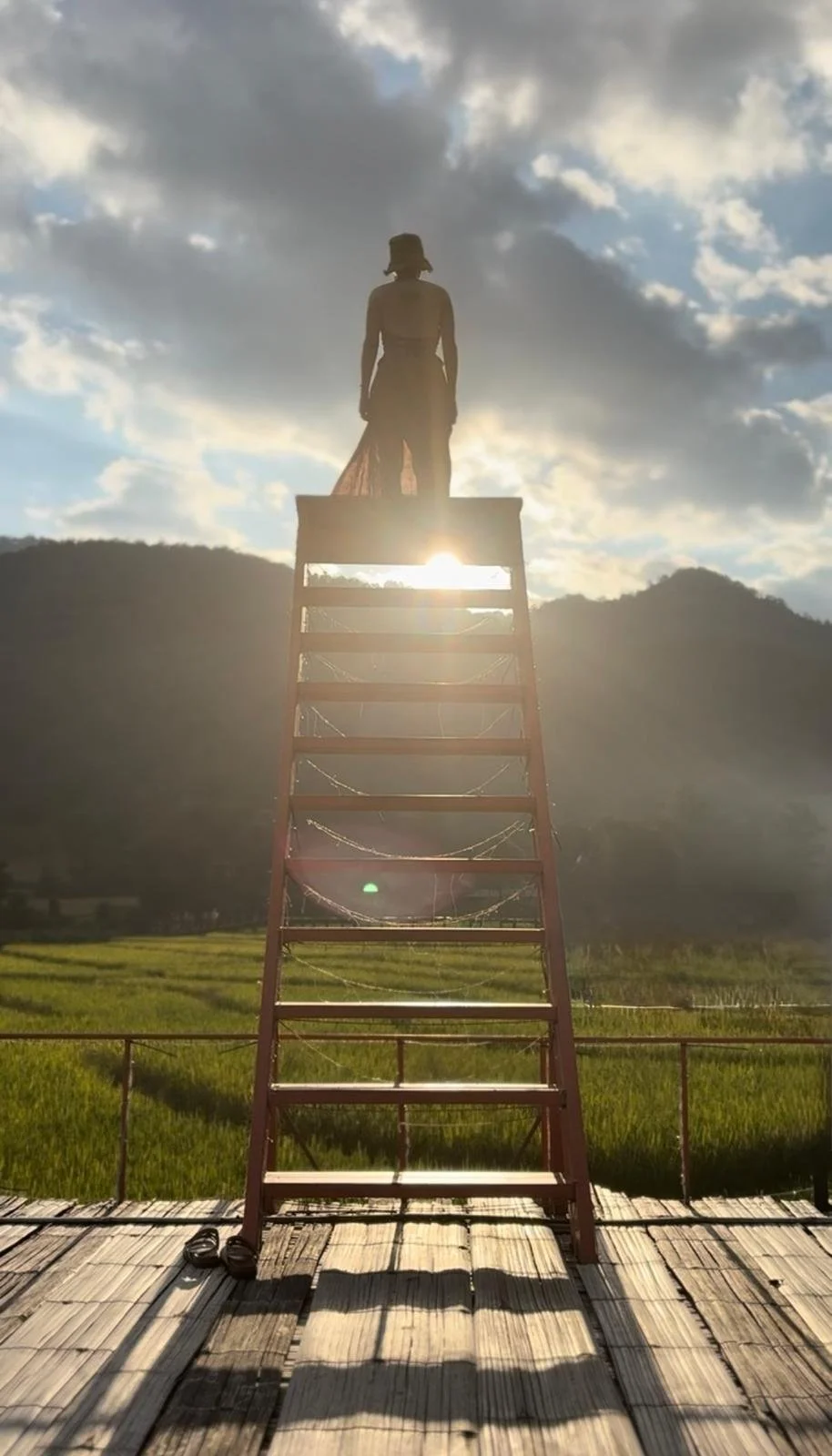 Une femme se tient au sommet d'une échelle en métal rouge, avec le soleil derrière elle, dans un paysage de foret et de rizière thailandaise, sous un ciel partiellement nuageux.