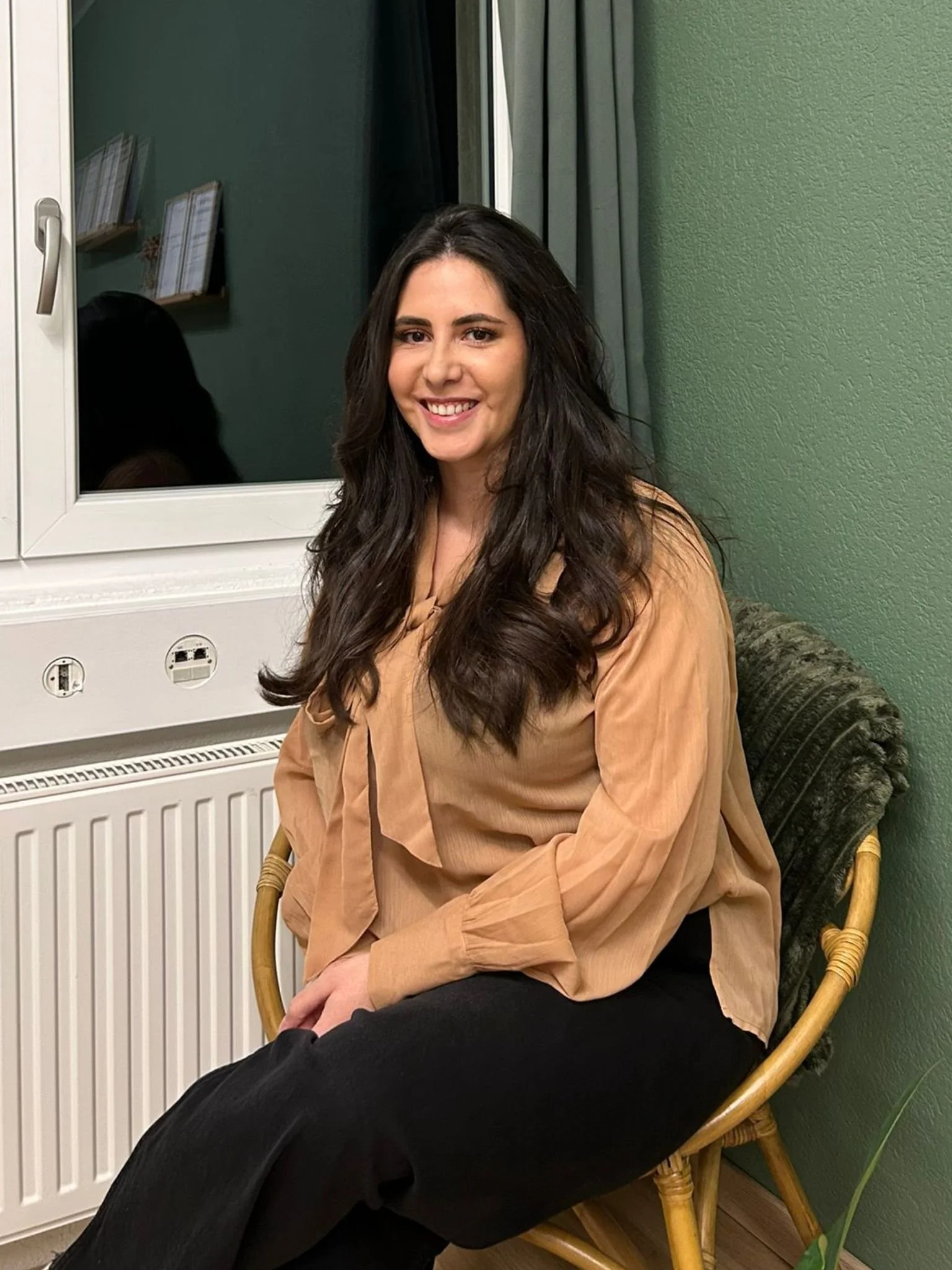 Une femme avec de longs cheveux bruns assise sur une chaise en rotin, souriante, portant un chemisier beige et un pantalon noir, dans une pièce verte avec un mur à gauche et une fenêtre derrière elle.