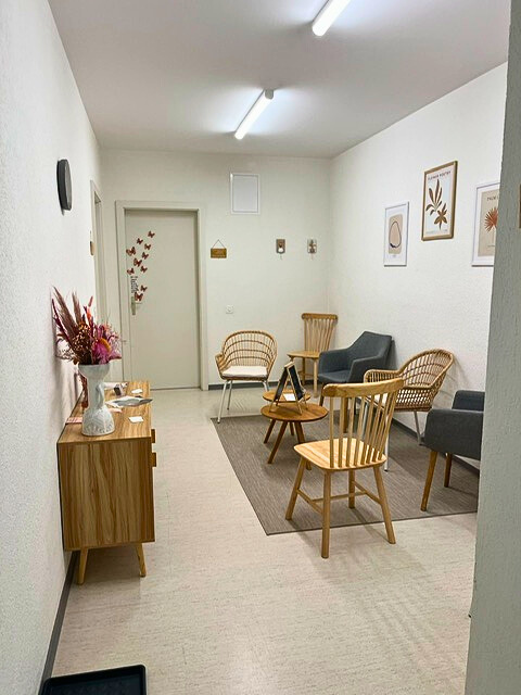 Salle d'attente espace détente, chaises en bois et mobilier calme et relaxant, décorations florales, des tableaux modernes sur le mur, et une porte au fond. Lampes au plafond