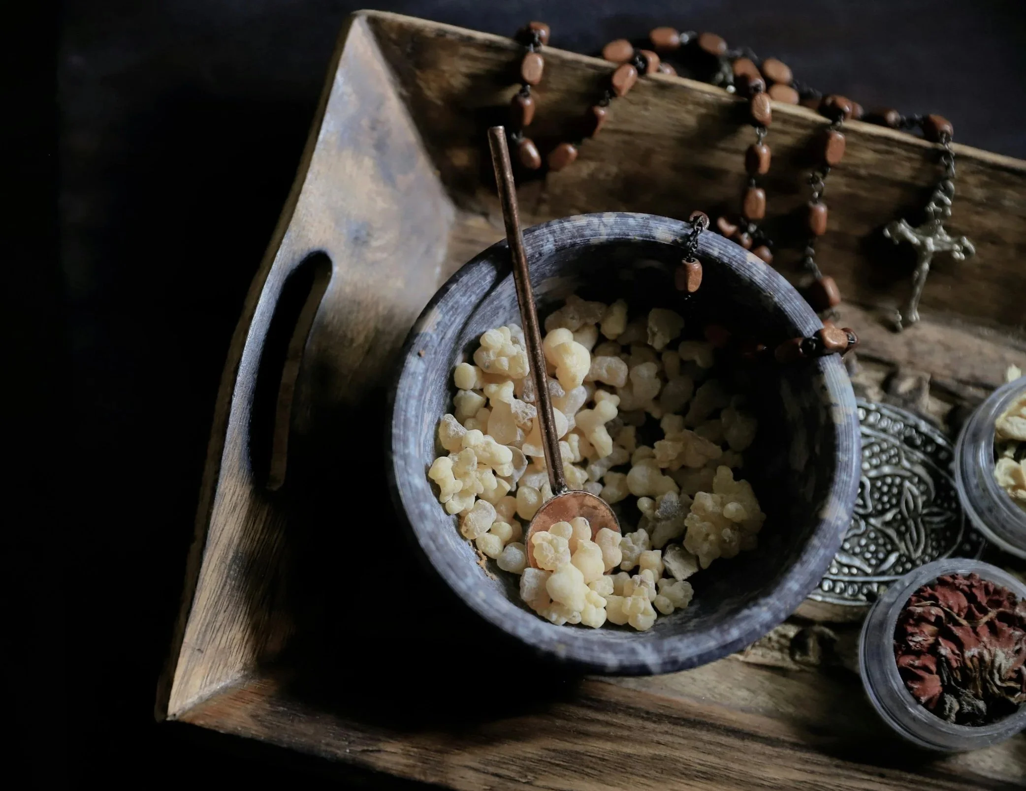 Close-up of a tabletop with various traditional medications, including a bowl of yellowish pills, a rosary, and other small bowls filled with herbs and dried ingredients.