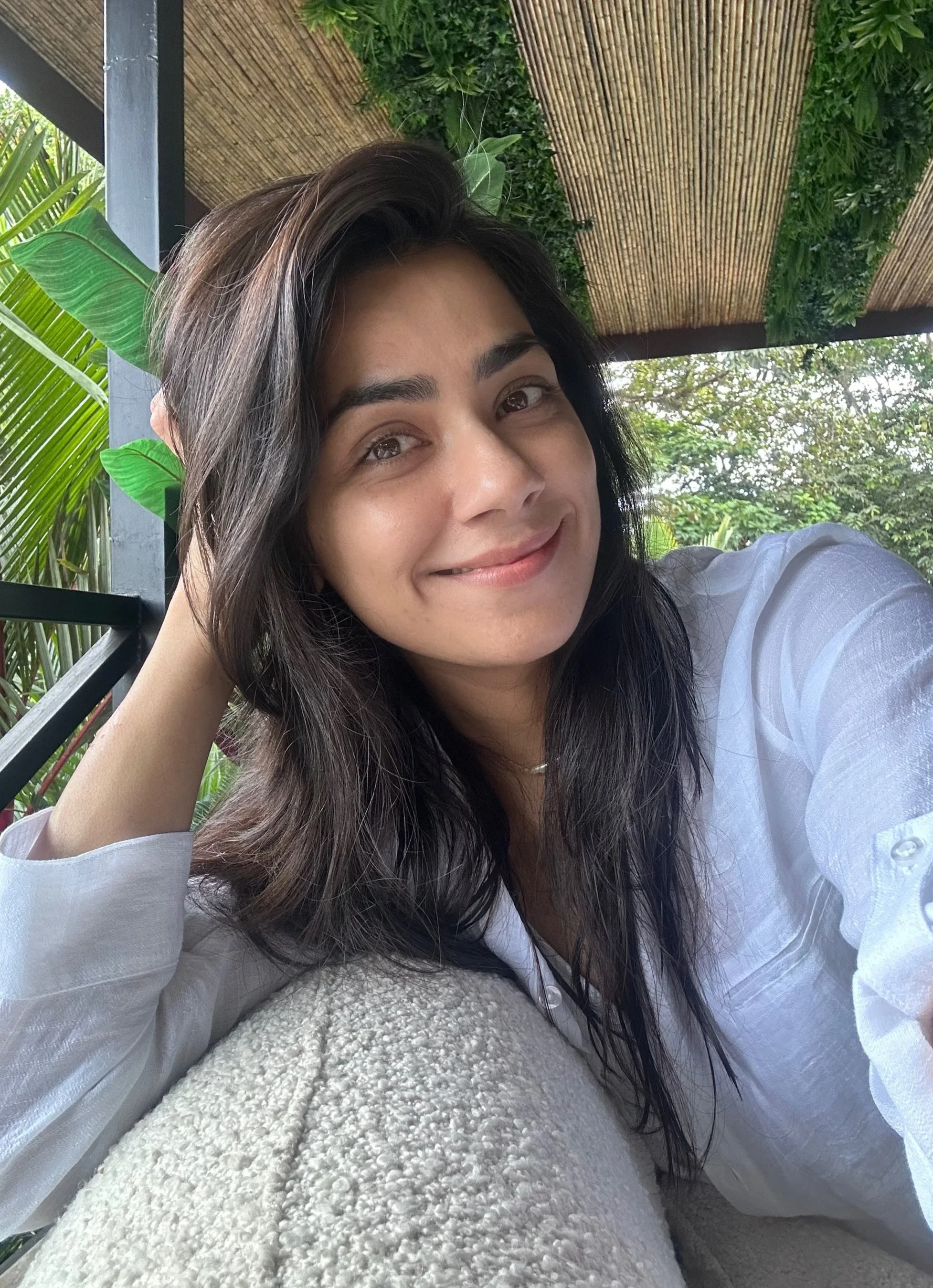 A woman with long dark hair, wearing a white shirt, smiling and resting her head on her hand at an outdoor setting with green plants and a bamboo roof in the background.