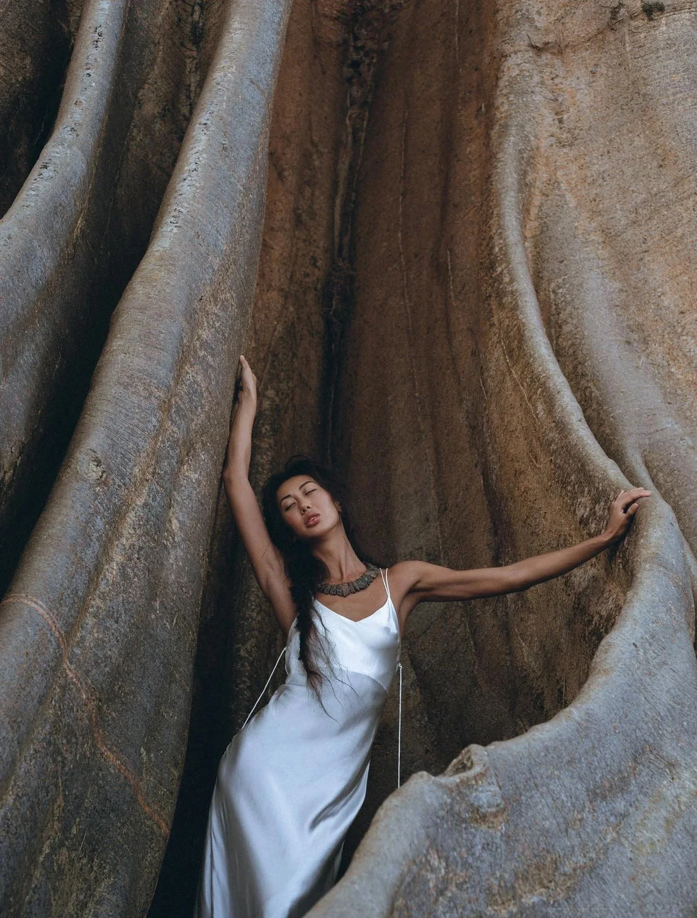 A woman with long dark hair and light skin, wearing a white dress and a statement necklace, posing with her arms raised and her back against a large natural rock formation.