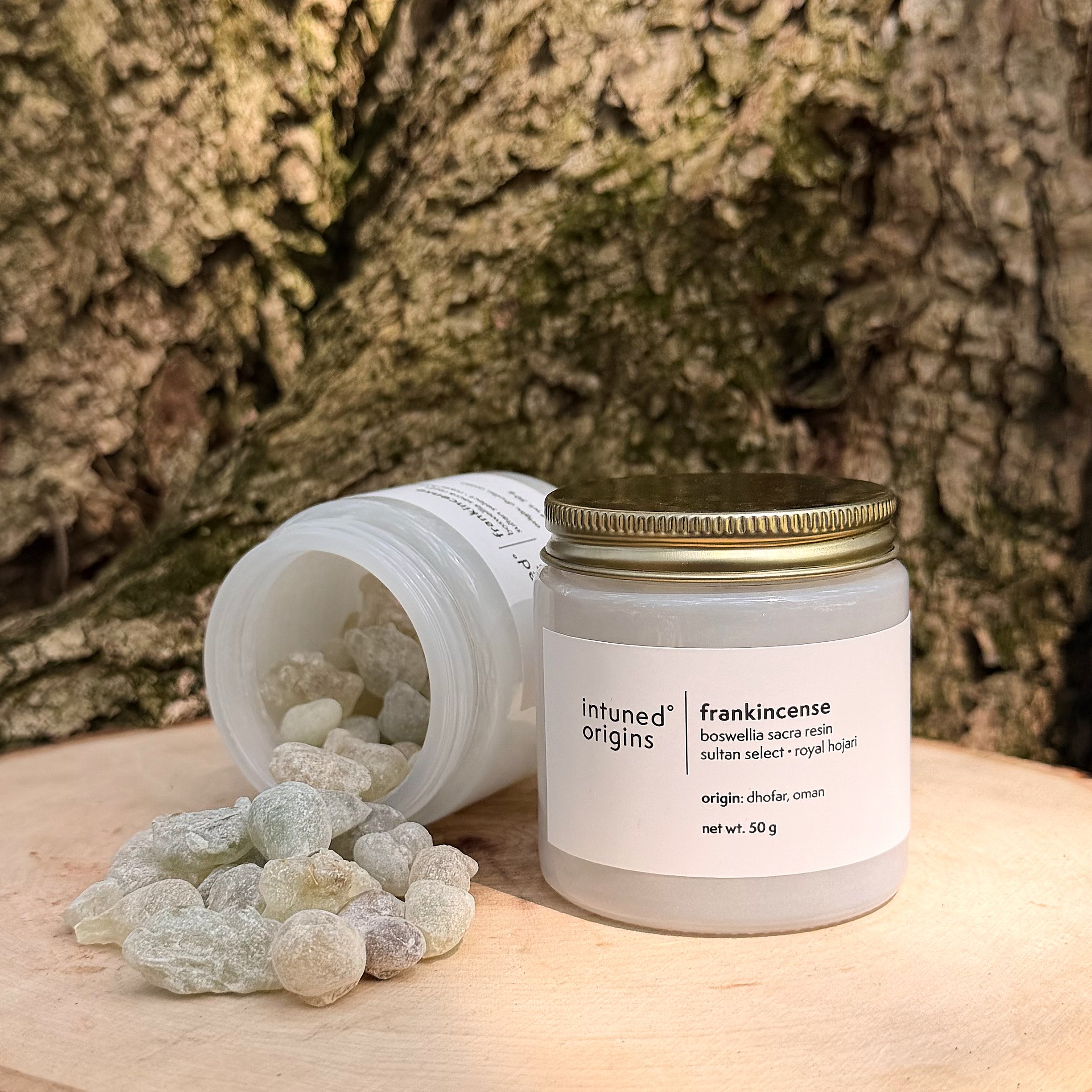 Close-up of a white container with a gold lid, labeled 'intuned origins' and 'frankincense,' placed on a light wooden surface. The container is surrounded by small, rounded, pale gray and greenish stones. The background features a rough, brown, textured surface.