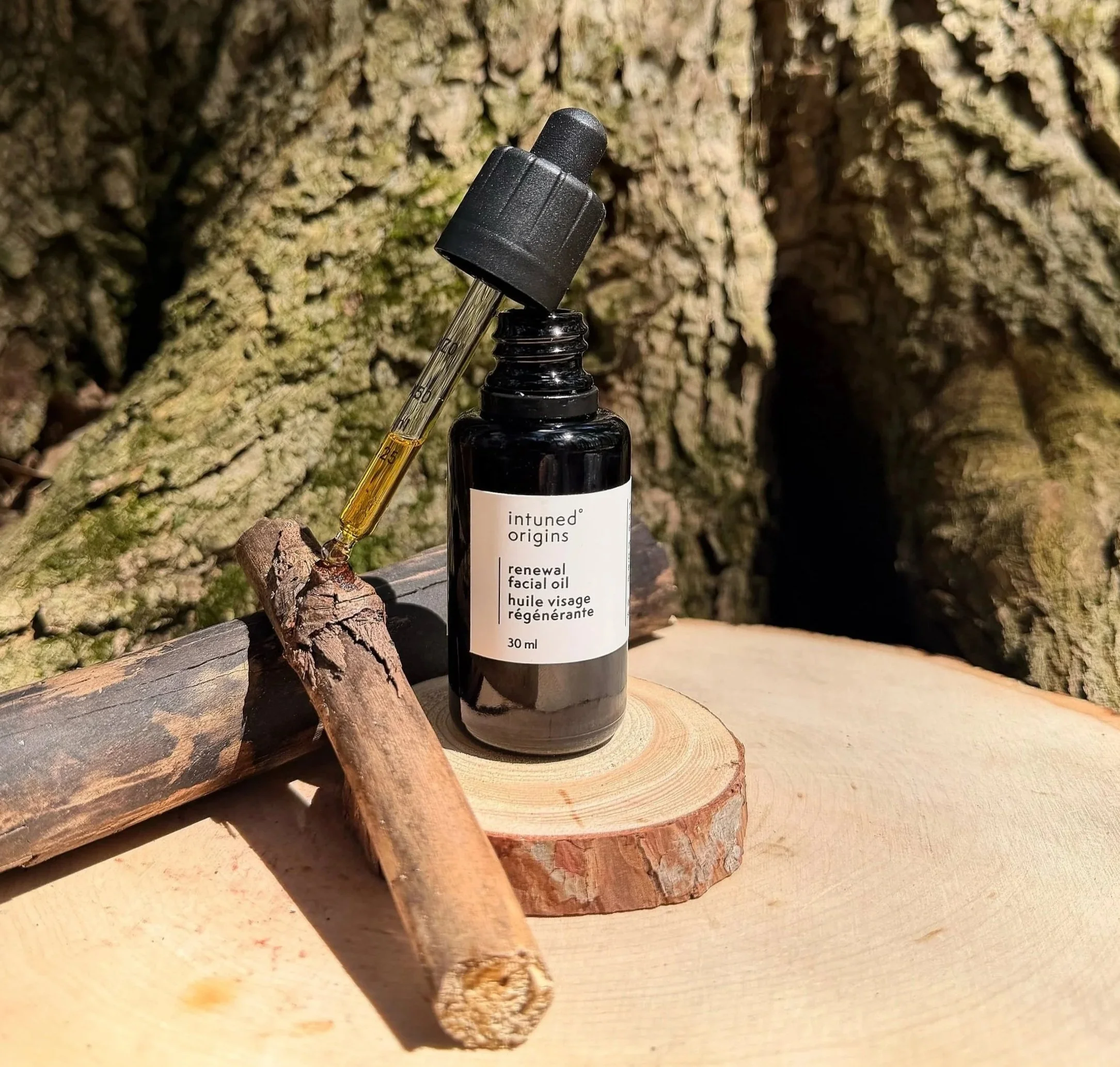 A small glass bottle labeled 'intuned origins renewal facial oil' with a black dropper cap, placed on a wooden surface, surrounded by pieces of wood and bark outdoors.