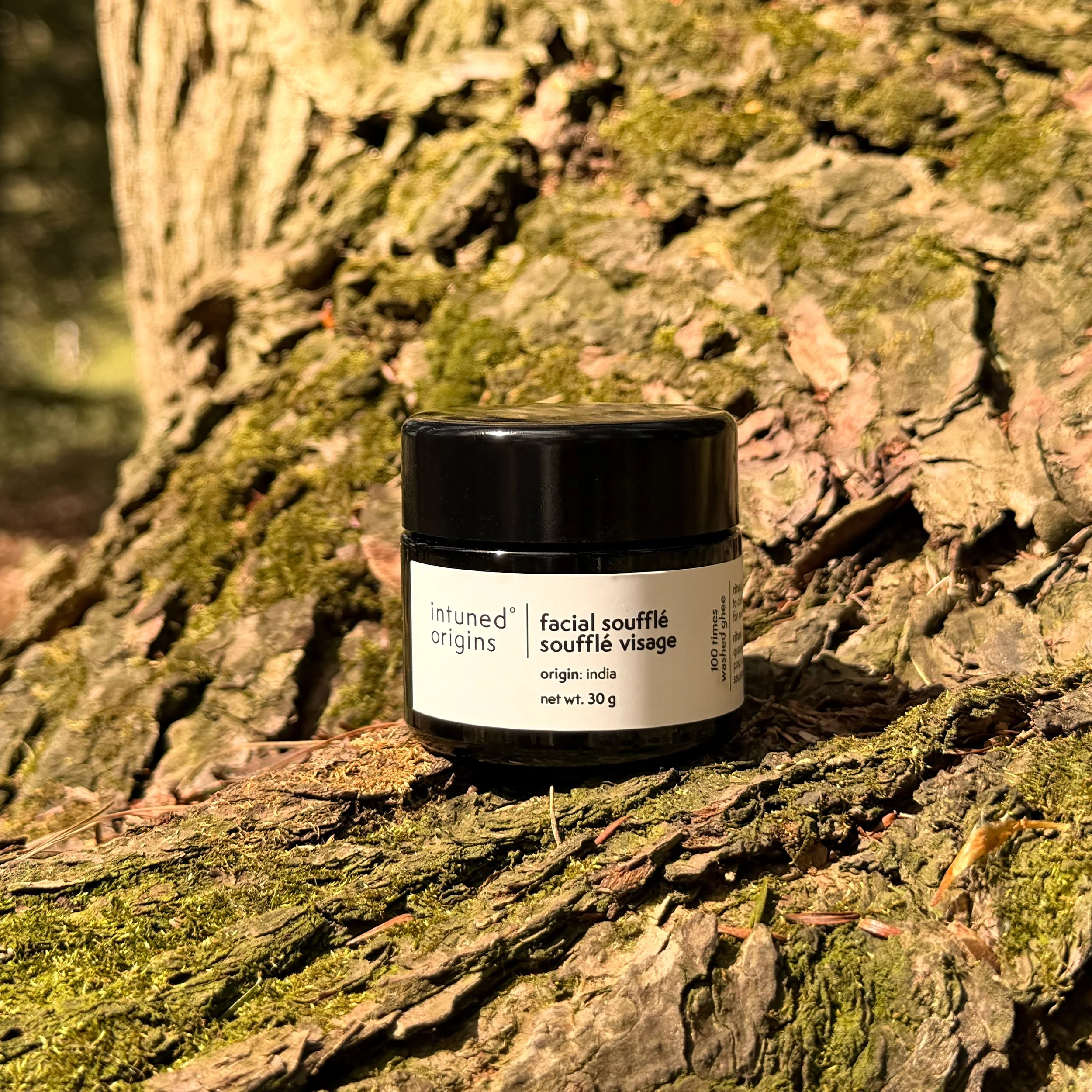 A black bottle of facial souffle cream placed on a tree trunk with rough bark and moss, outdoors.