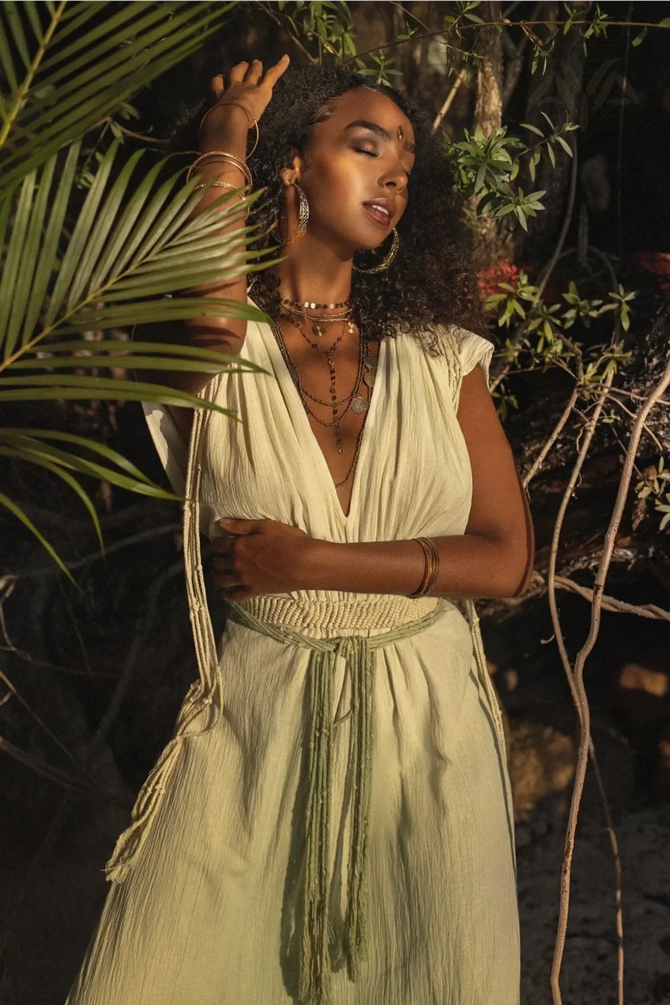 A woman with dark curly hair and brown skin, dressed in a light yellow, flowing dress with a tied waist, standing among green plants and branches at night with closed eyes and a serene expression.