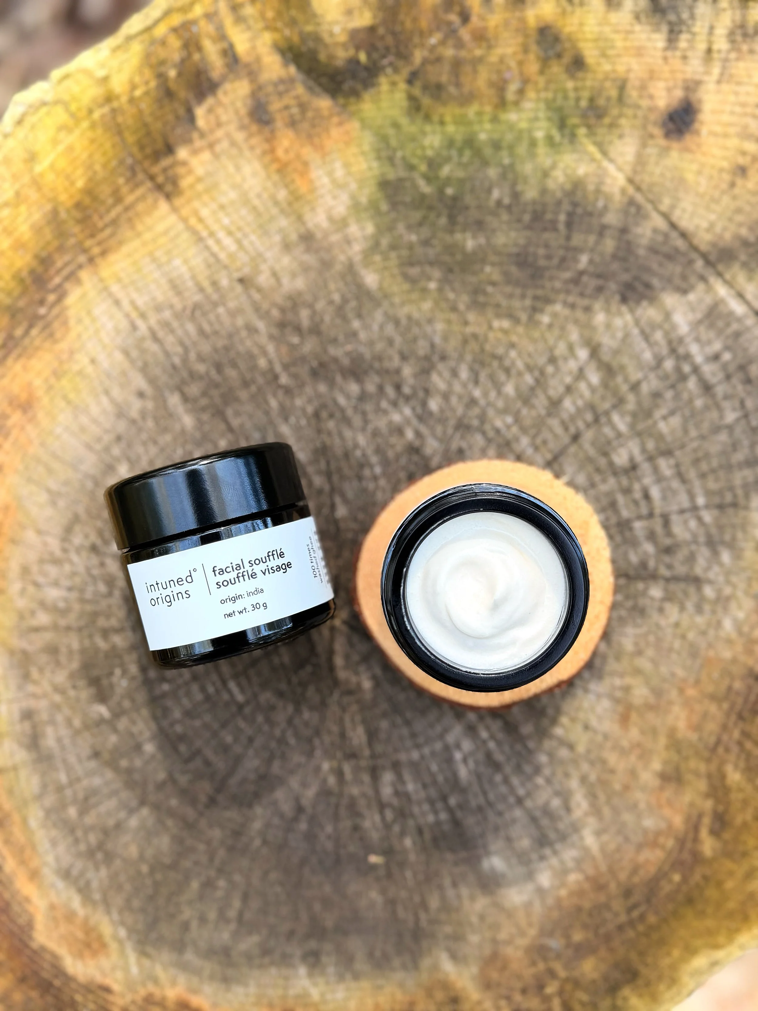 A black jar labeled 'Intuned Origins facial souffle' on a wooden surface, with an open jar of white cream next to it.