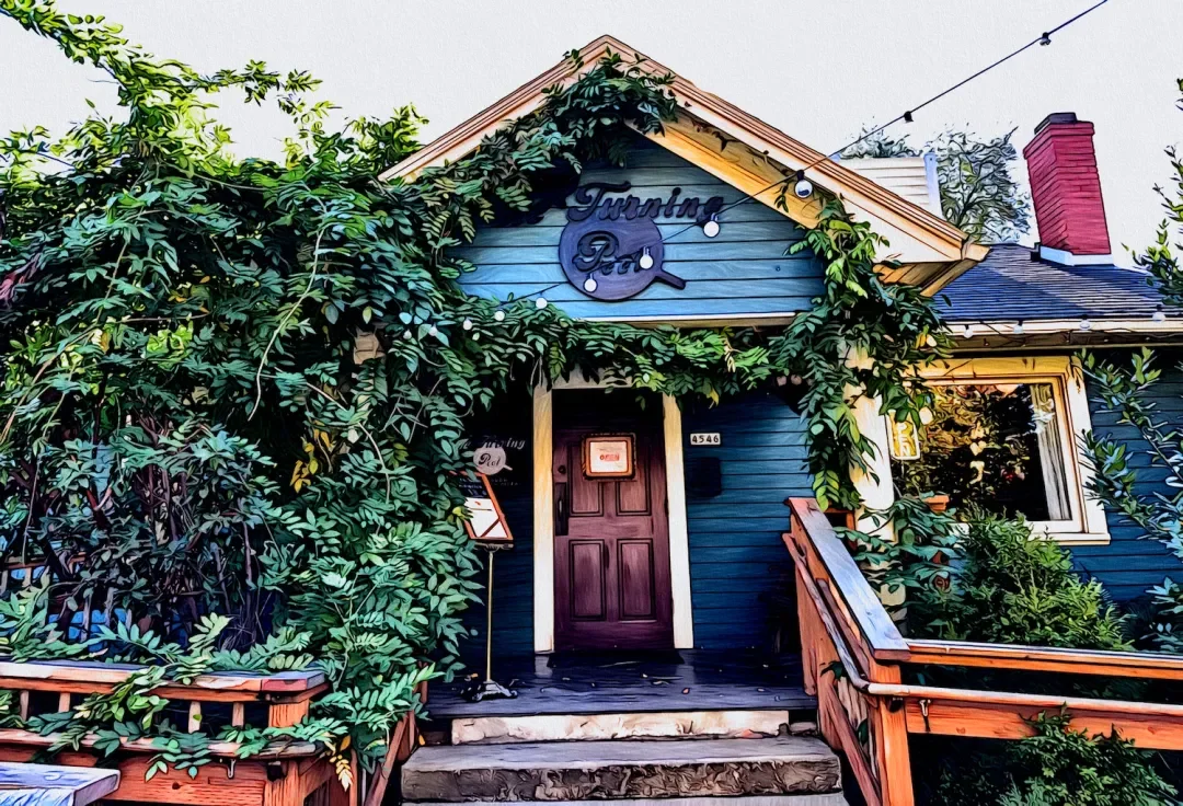 Blue house with a porch, surrounded by lush green vines and plants, with a 'Tasting Room' sign on top of the house, and string lights hanging overhead.
