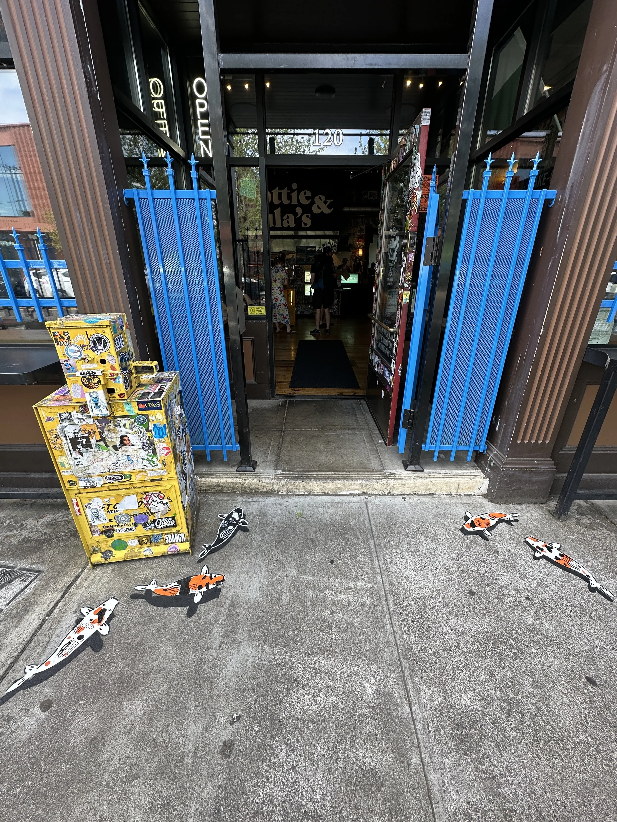 The entrance of a restaurant or cafe with a black door frame and a sign that says '120'. Blue vertical metal bars serve as a gate on each side of the door. Outside, there are decorative koi fish cutouts on the concrete sidewalk and a yellow vending machine covered in stickers on the left side.