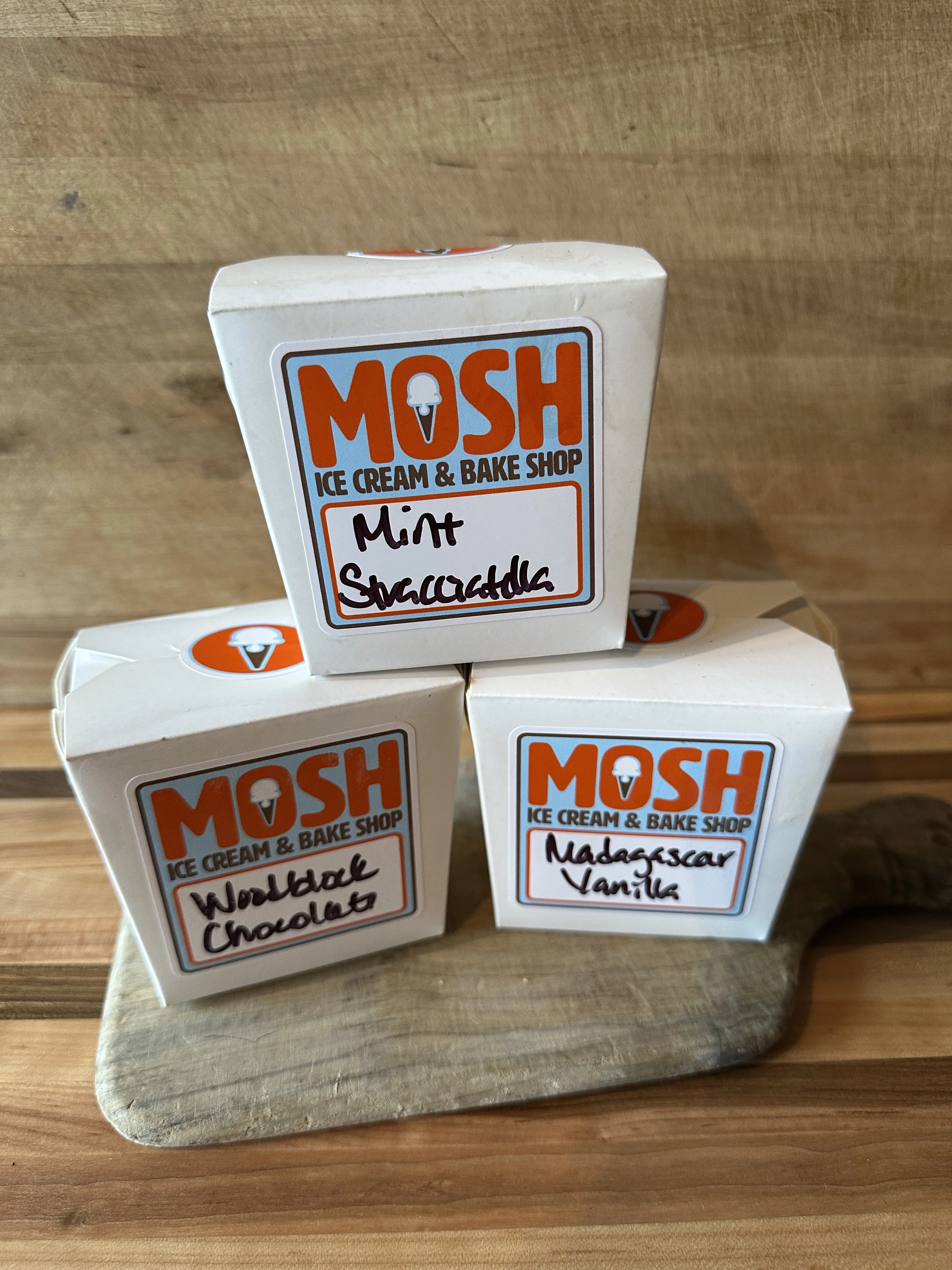 Three small ice cream cartons labeled 'Mosh Ice Cream & Bake Shop' with flavors 'Mint Chocolate,' 'Wondotal Chocolate,' and 'Madagascar Vanilla' placed on a wooden board against a wooden background.