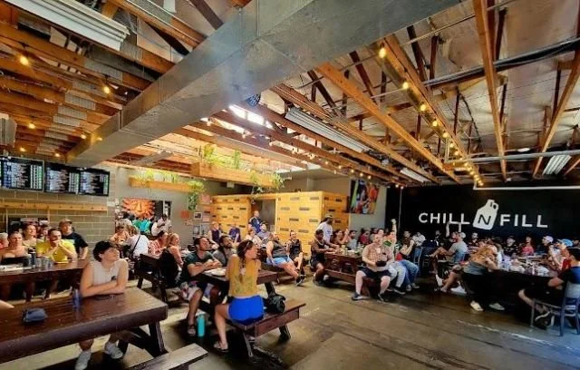 People sitting on benches and tables in a modern restaurant or café with wooden ceiling beams and exposed ductwork, and a black wall with the sign 'CHILL N FILL' in the background.