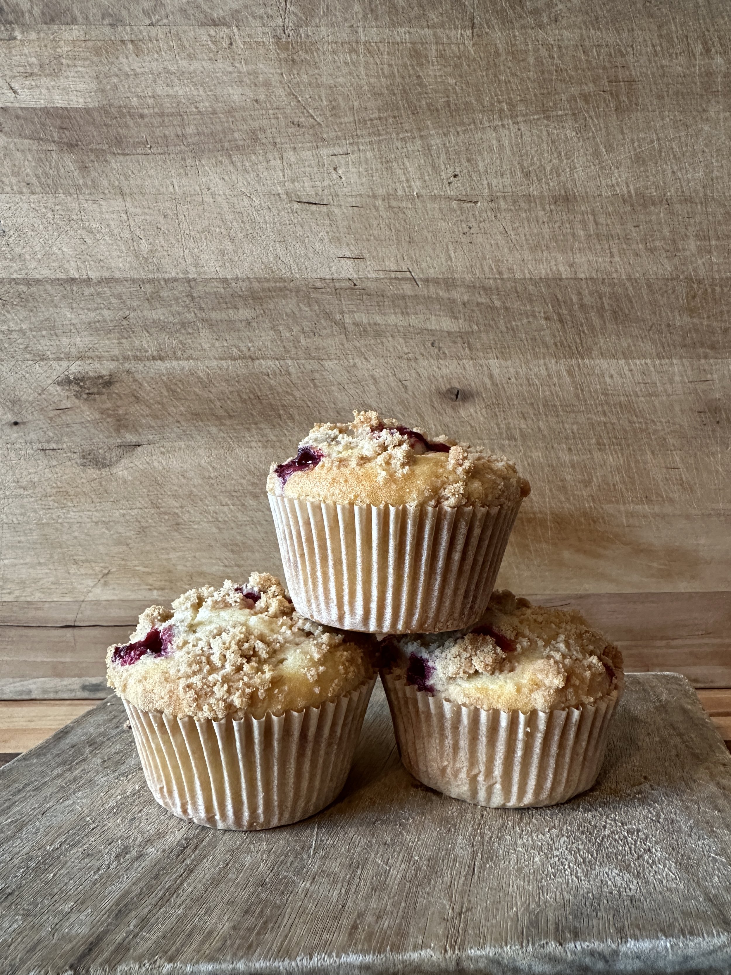 Three blueberry muffins with crumb topping arranged in a pyramid on a wooden surface against a wooden background.