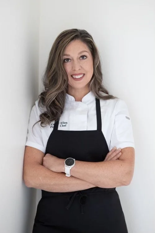 A woman with long wavy brown hair smiling, wearing a white chef's coat with name tag, black apron, and a white watch, standing with arms crossed against a white wall.