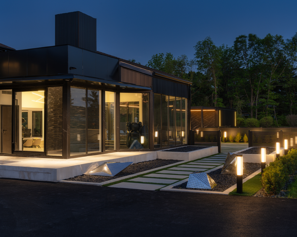 Modern house with large glass windows and a landscaped yard illuminated at night, featuring pathway lights and decorative outdoor fixtures.