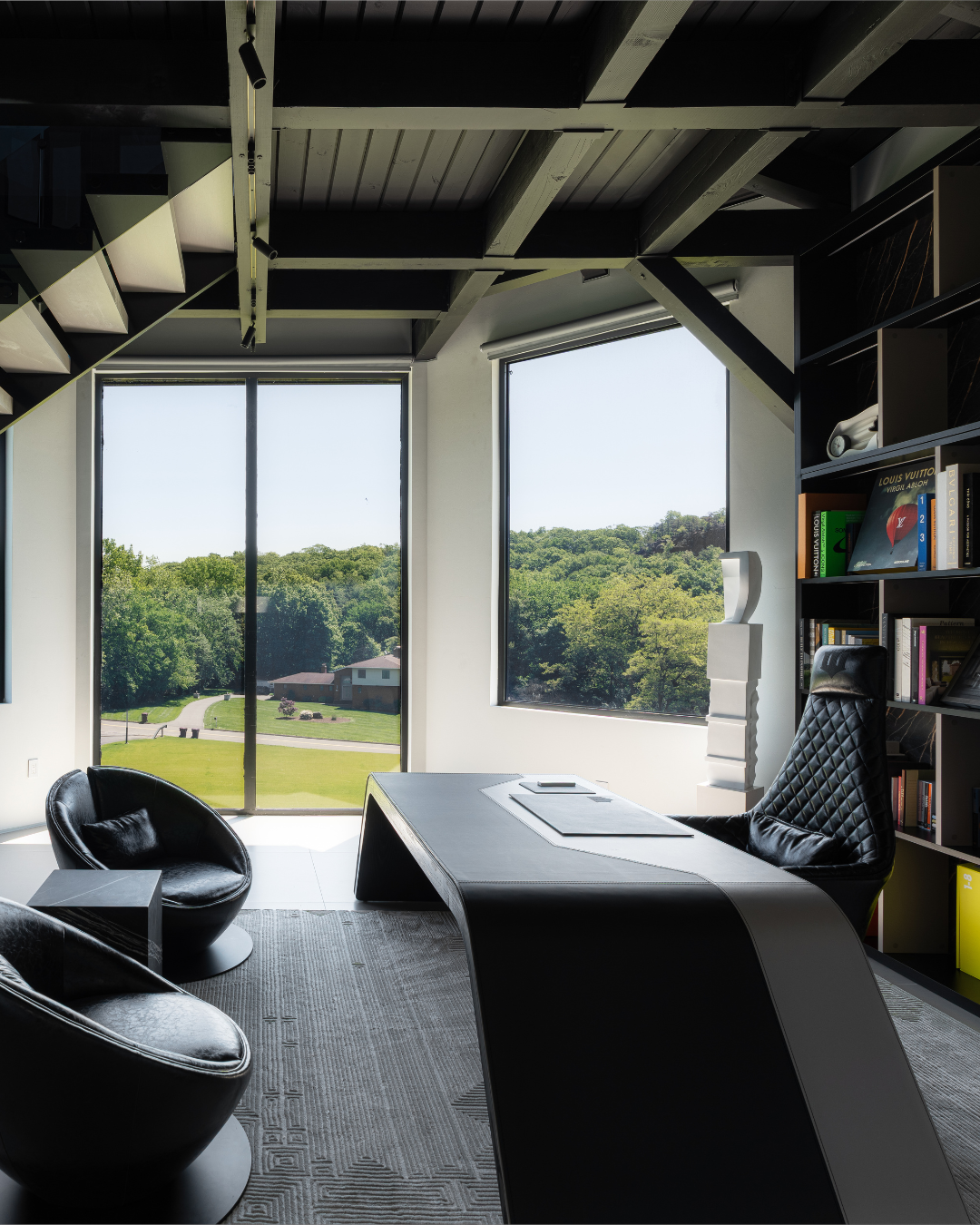 Modern office with large windows overlooking green landscape, black leather chair, bookshelf, and contemporary decor.