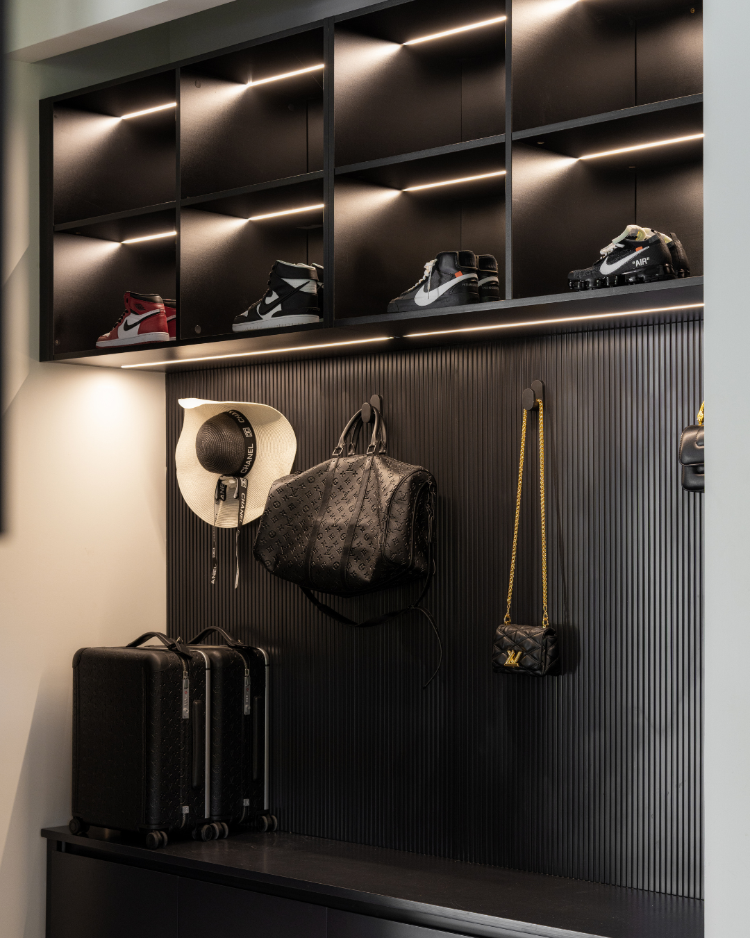 Display of luxury sneakers, handbags, a hat, and luggage on a black wall with modern lighting.