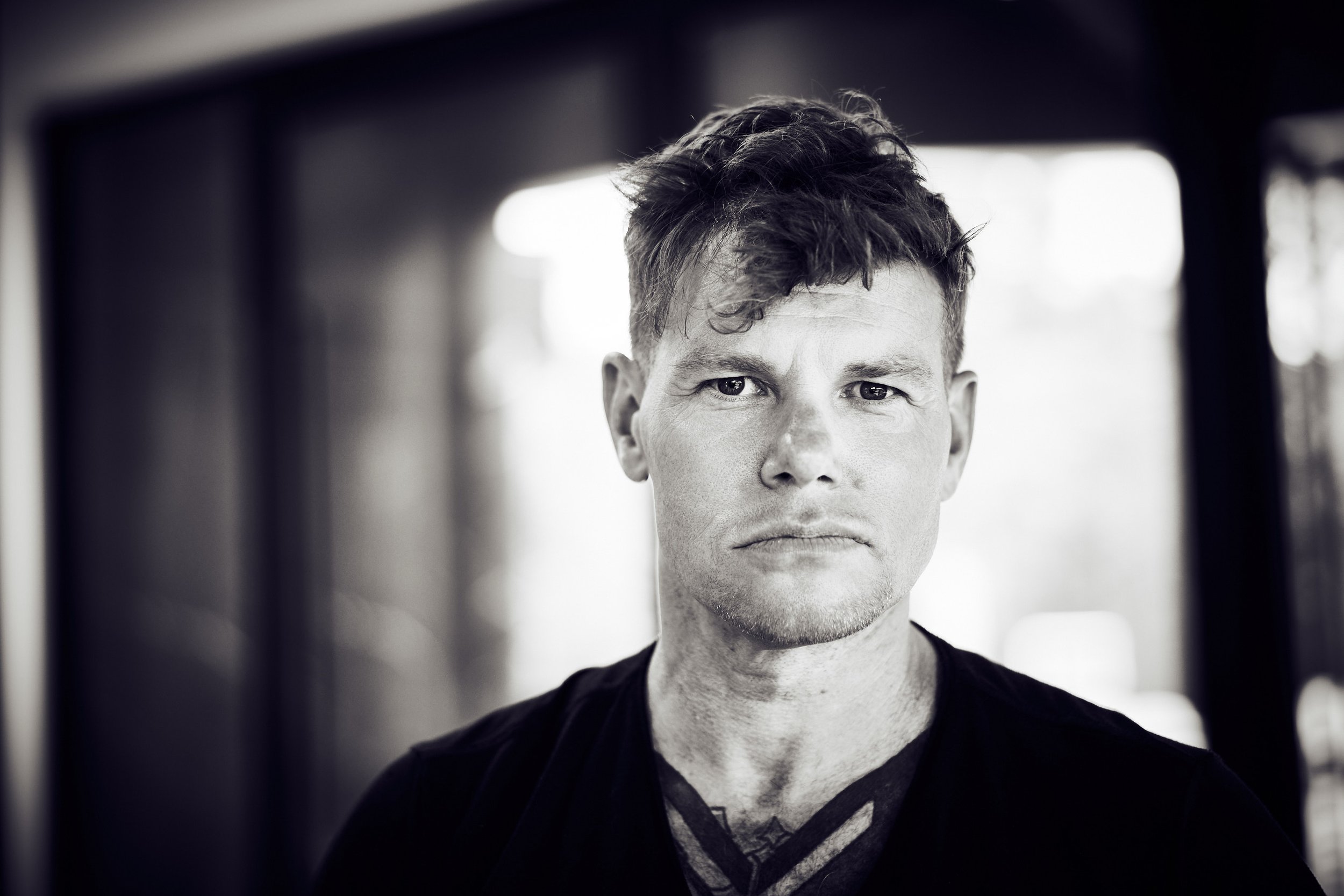 A black and white portrait of a young man with short, tousled hair, wearing a dark shirt, looking directly at the camera with a serious expression.