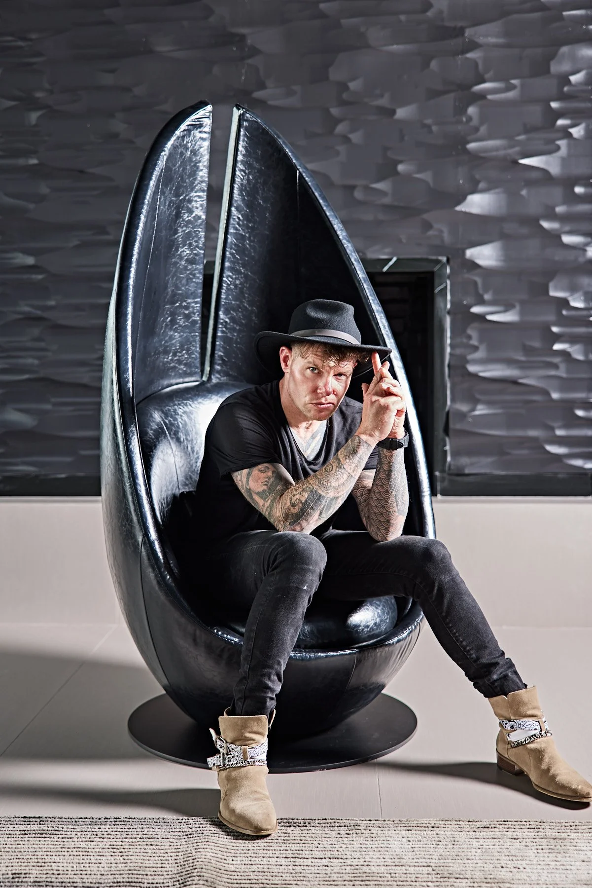 A man sitting in a black egg-shaped chair, wearing a black hat, black T-shirt, black jeans, and tan boots with decorative straps, looking at the camera with a serious expression. Background features a textured black wall.