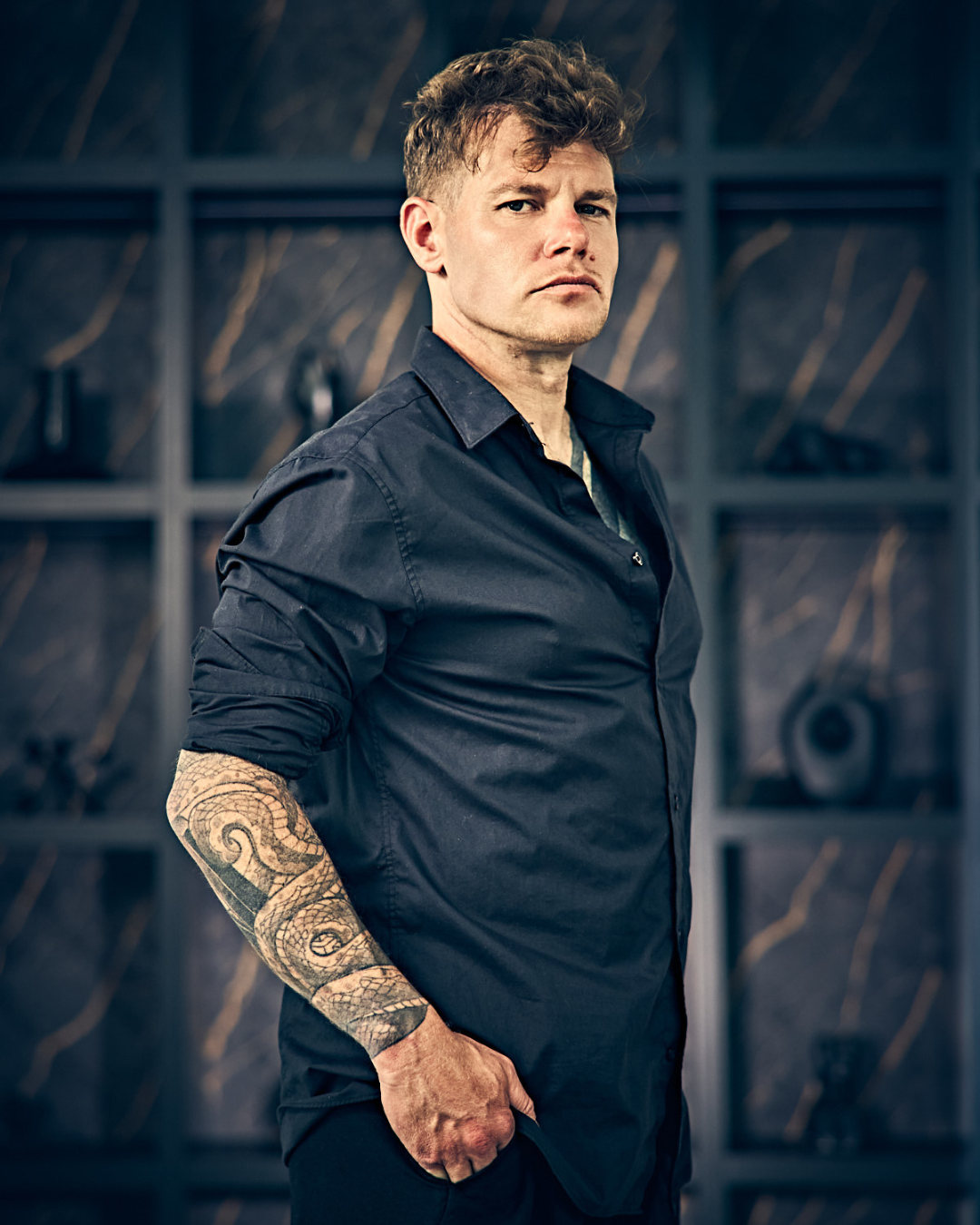 A man with a tattooed arm wearing a dark blue shirt, standing in front of a dark wall with shelves of swords and historical weapons.