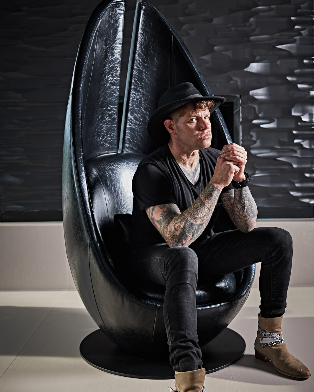 A man with tattoos wearing a black hat, black T-shirt, and jeans, sitting in a black egg-shaped chair with a modern dark interior background.