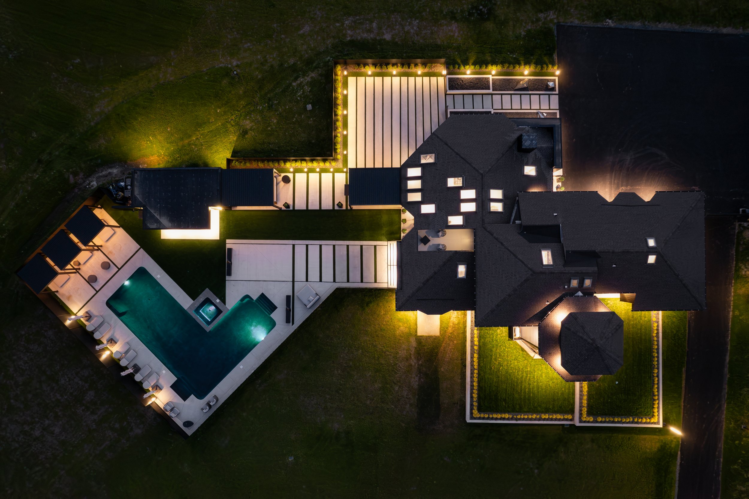 Aerial view of a modern house at night with outdoor lighting, a swimming pool, patio area, and landscaped yard.