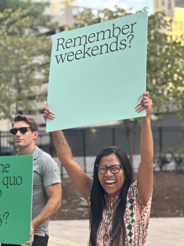 Woman holding a light blue protest sign that says "Remember weekends?" at an outdoor event.
