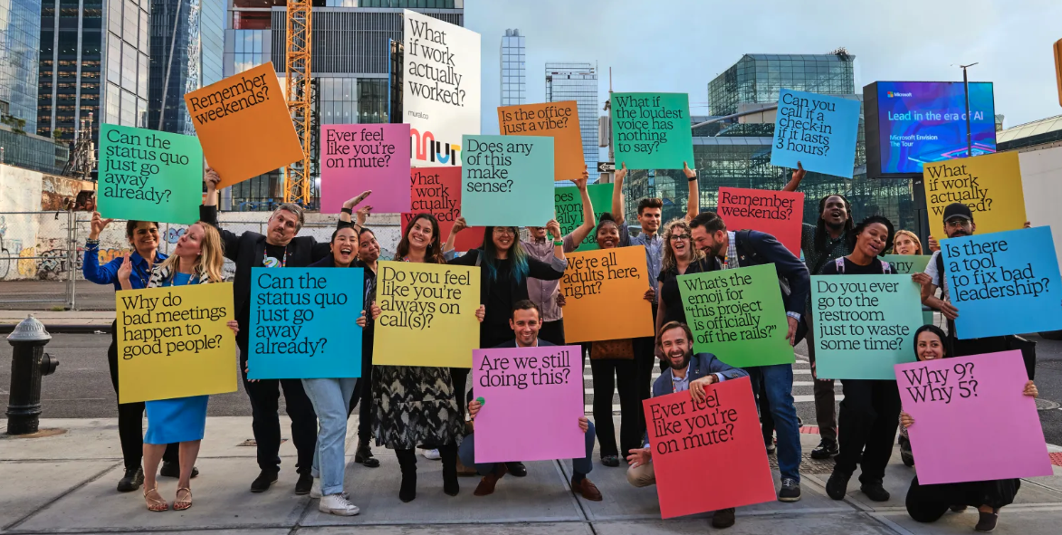 A diverse group of people holding colorful protest signs with questions and statements about work and leadership, standing outdoors in an urban setting.