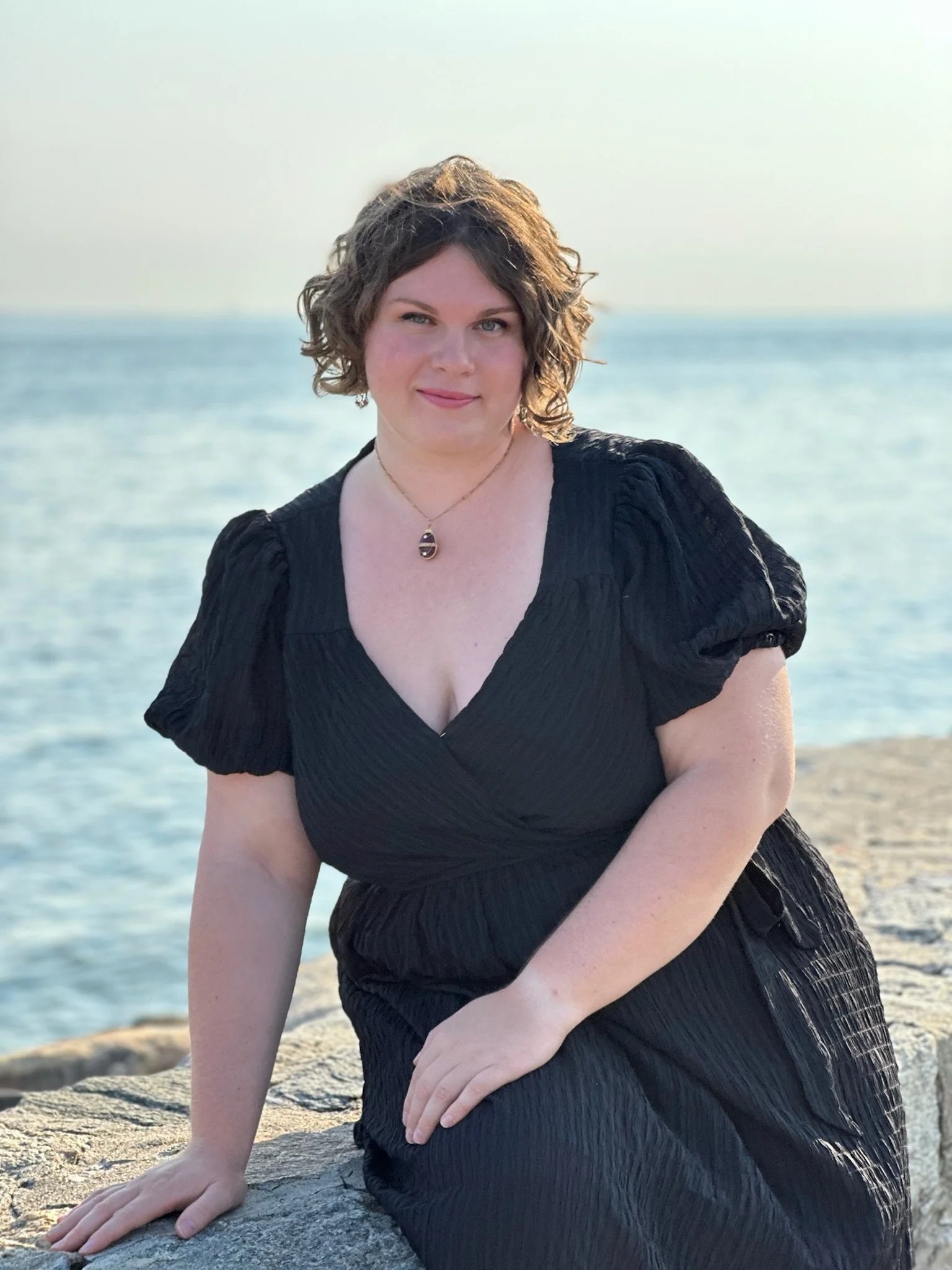 Sara Lynn Hargrave (Soprano) in a black dress sitting on rocks by the water at sunset.
