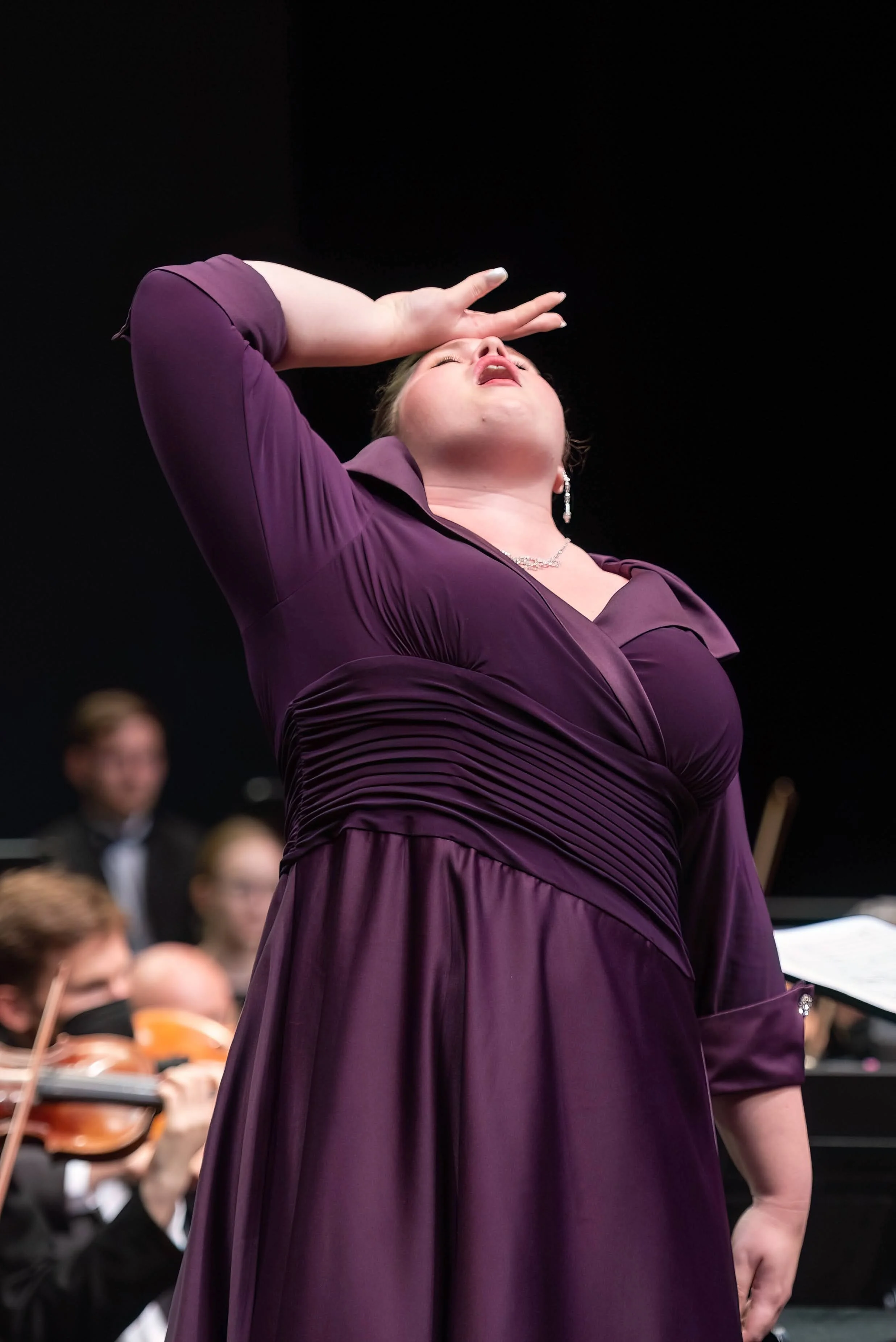 Sara Lynn Hargrave (Soprano) in a purple dress is performing passionately with her eyes closed and hand on her forehead, while orchestral musicians play in the background.