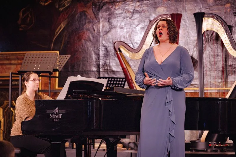 Sara Lynn Hargrave (Soprano) in a blue dress singing on stage with a pianist playing a black grand piano beside her at Minoritensaal in Graz.